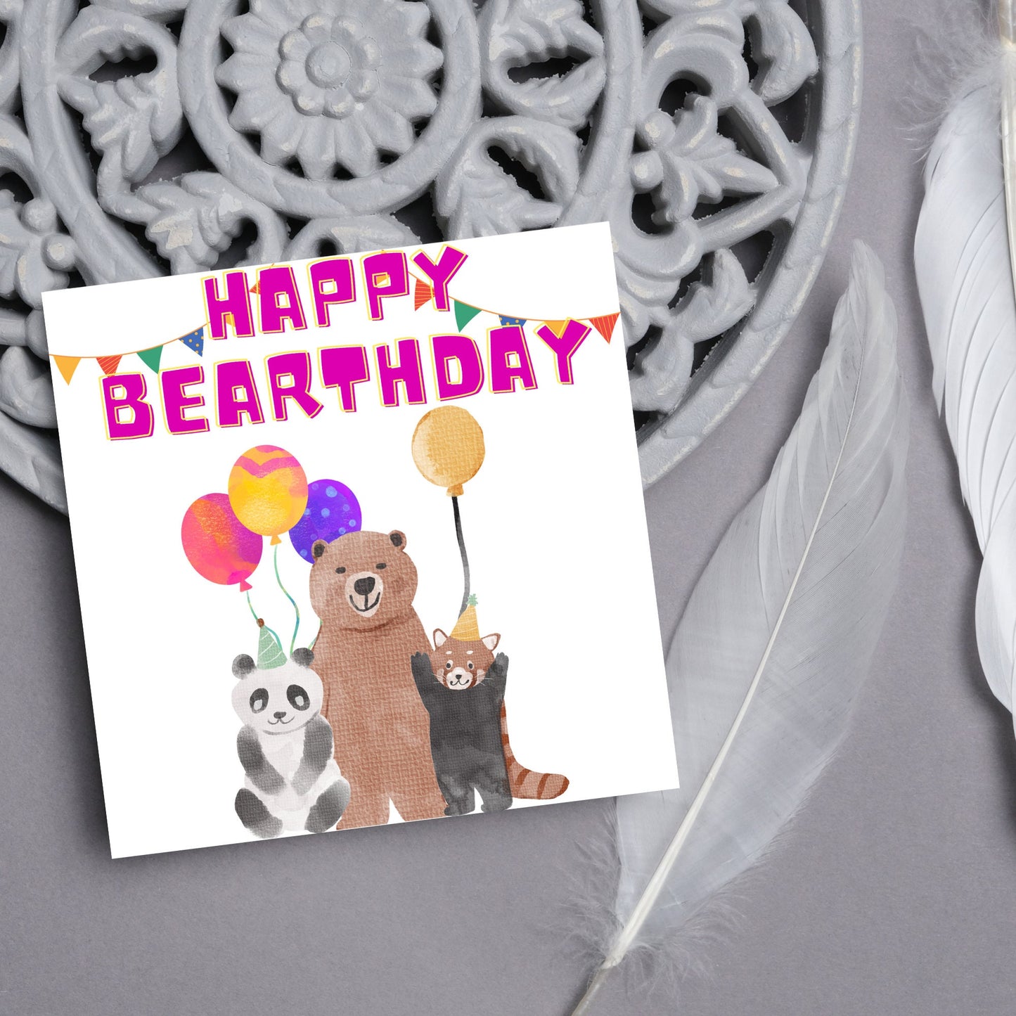 Image of a square Birthday Card. The card has a white background with a vibrant watercolour illustration of 3 bears holding balloons and big pink text that reads happy bearthday, a cute play on the traditional happy birthday.
