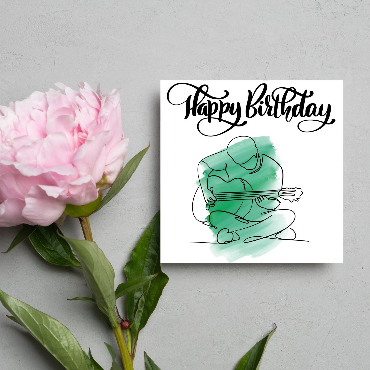 Square birthday card featuring a line drawing of a person sitting cross legged playing a guitar with a green watercolour style backdrop. The top of the card reads Happy Birthday in black swirly writing