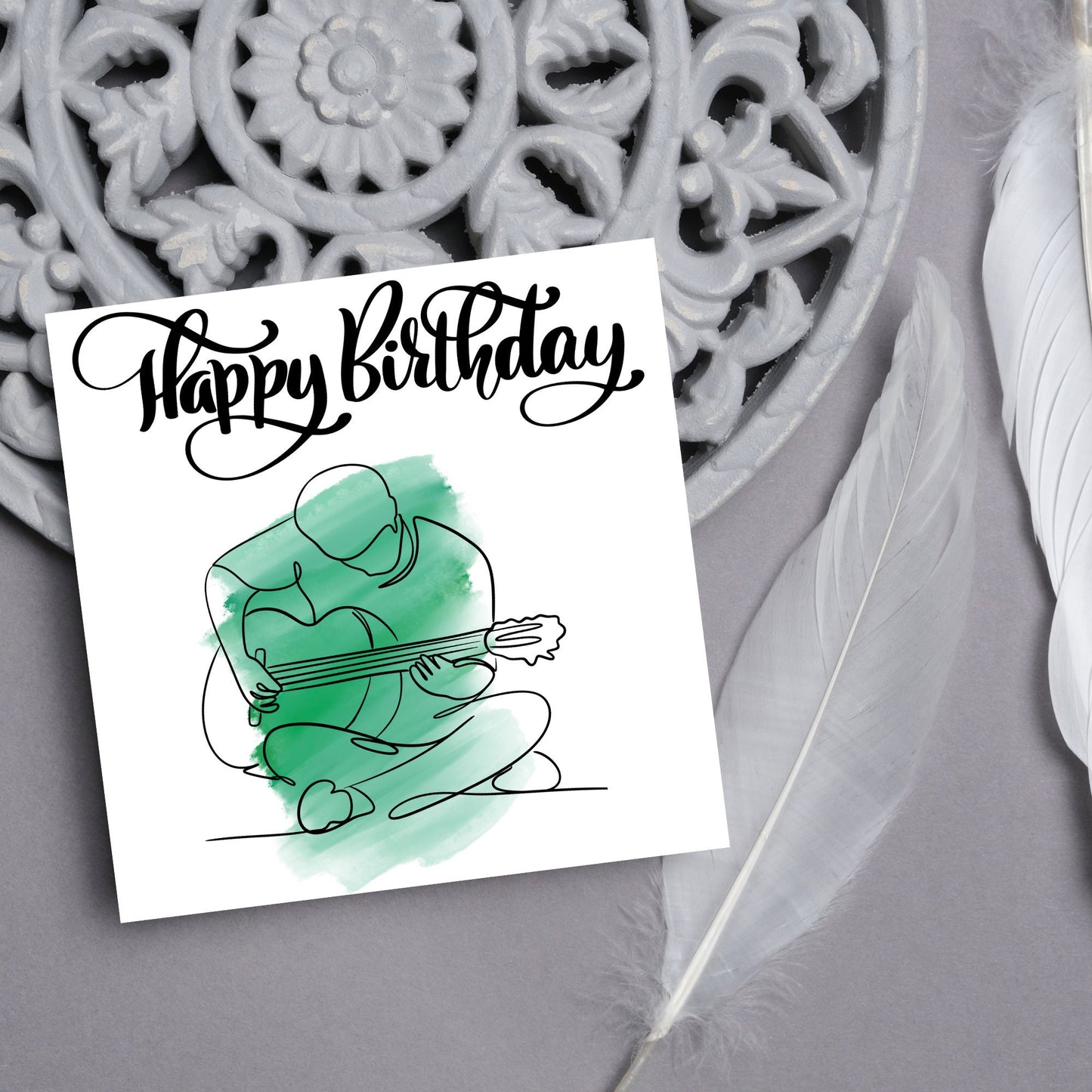 Square birthday card featuring a line drawing of a person sitting cross legged playing a guitar with a green watercolour style backdrop. The top of the card reads Happy Birthday in black swirly writing