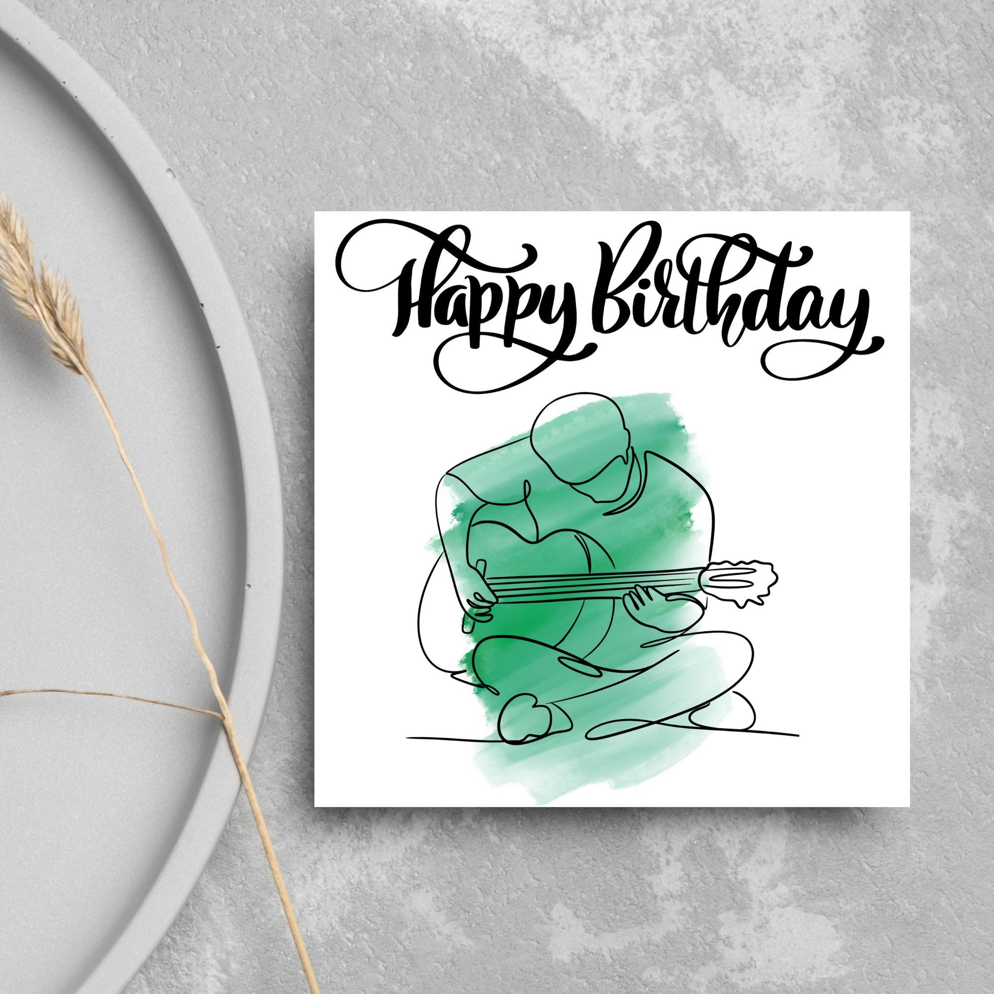 Square birthday card featuring a line drawing of a person sitting cross legged playing a guitar with a green watercolour style backdrop. The top of the card reads Happy Birthday in black swirly writing