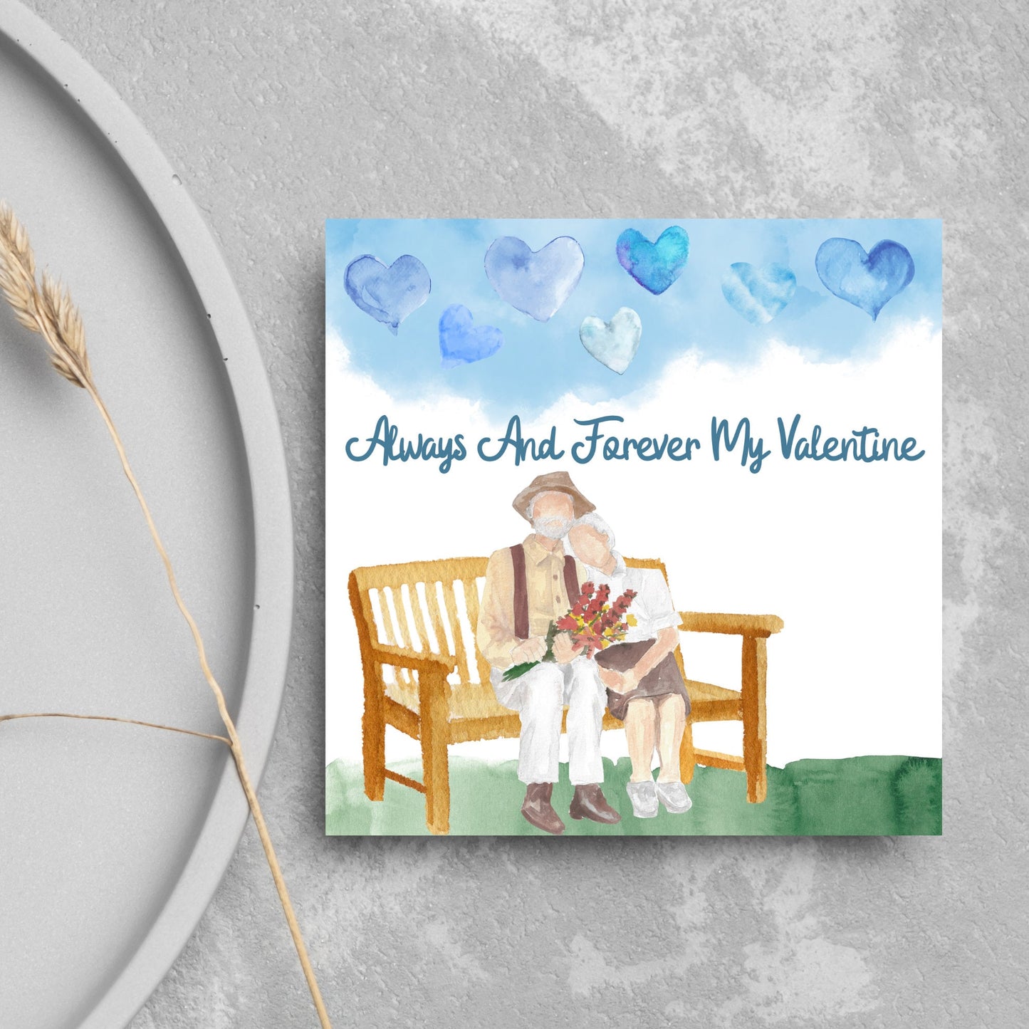 This beautiful Card has a watercolour style print with a couple sitting on a bench with flowers. Above them the blue sky has a series of cute watercolour hearts. This Cute card is perfect for valentines day for your wife or husband