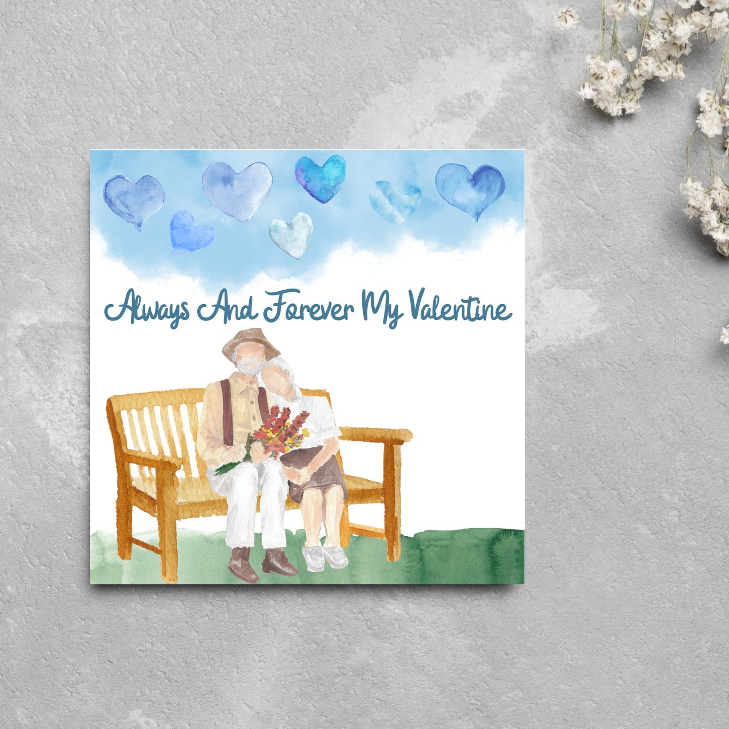 This beautiful Card has a watercolour style print with a couple sitting on a bench with flowers. Above them the blue sky has a series of cute watercolour hearts. This Cute card is perfect for valentines day for your wife or husband