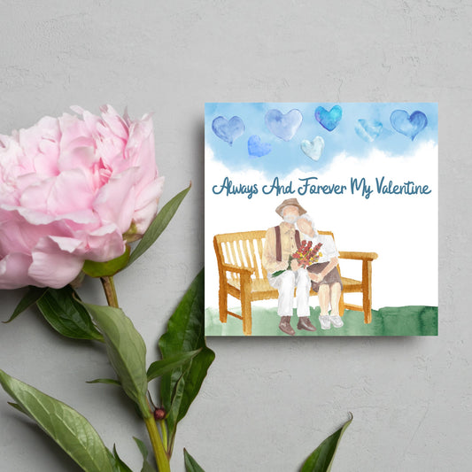 This beautiful Card has a watercolour style print with a couple sitting on a bench with flowers. Above them the blue sky has a series of cute watercolour hearts. This Cute card is perfect for valentines day for your wife or husband