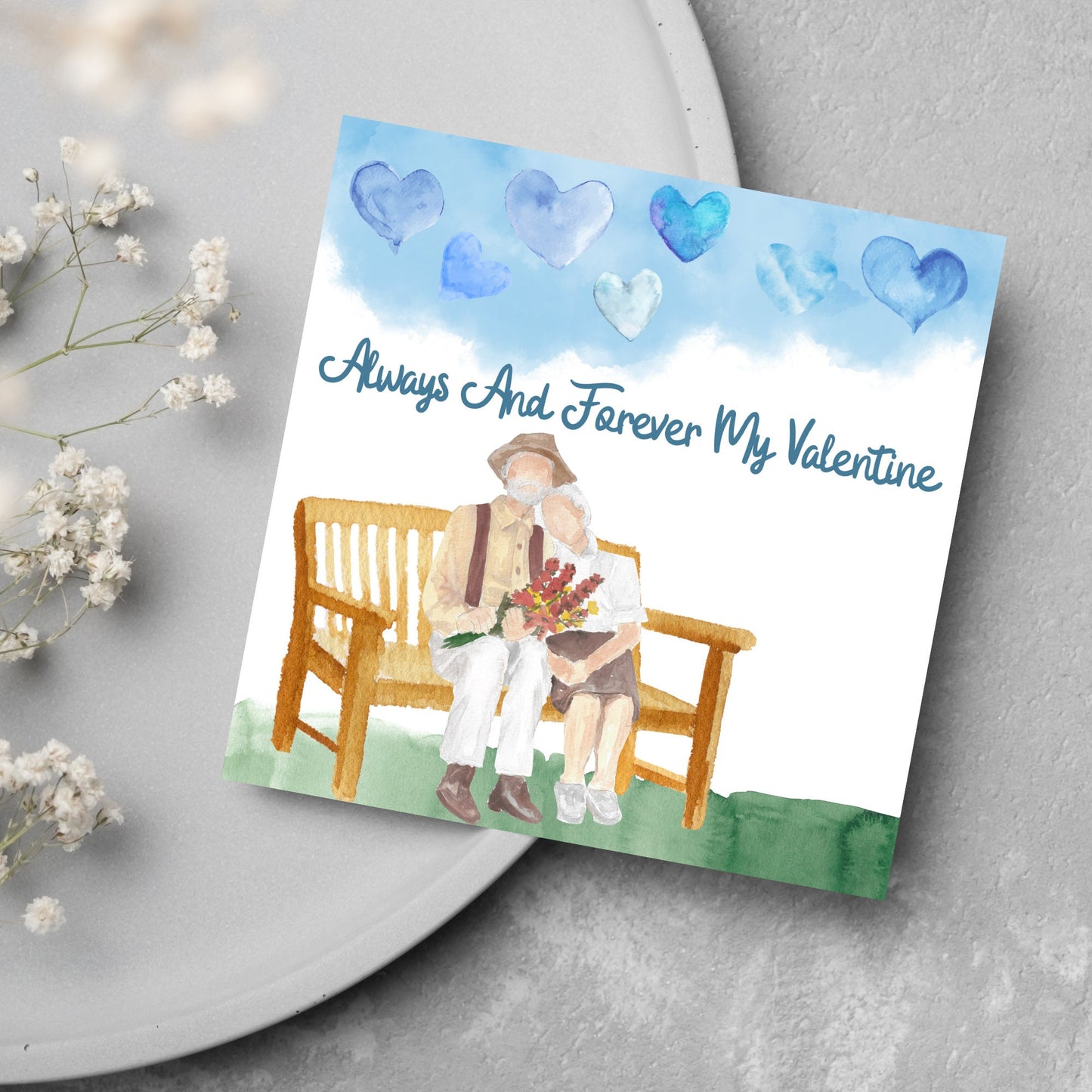 This beautiful Card has a watercolour style print with a couple sitting on a bench with flowers. Above them the blue sky has a series of cute watercolour hearts. This Cute card is perfect for valentines day for your wife or husband