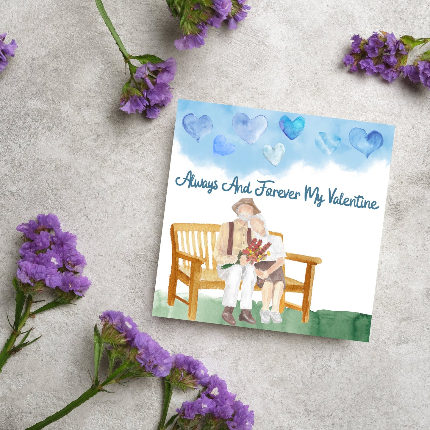 This beautiful Card has a watercolour style print with a couple sitting on a bench with flowers. Above them the blue sky has a series of cute watercolour hearts. This Cute card is perfect for valentines day for your wife or husband