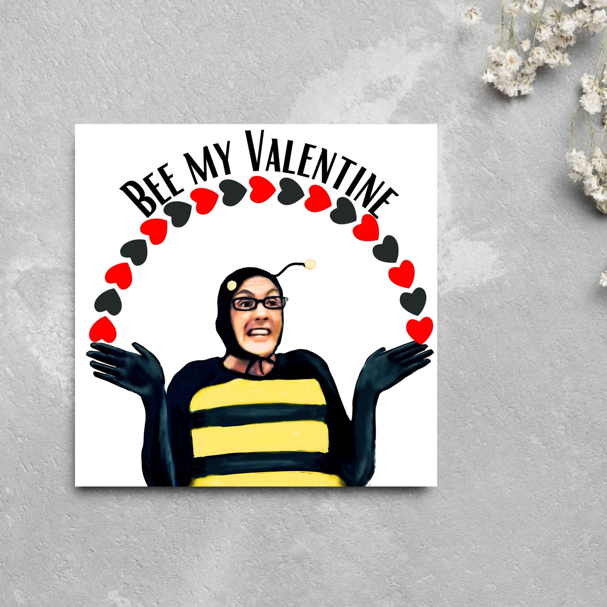 Square greetings card featuring Dean Pelton from the TV show Community, in one of his eccentric costumes dressed as a bee holding an arch of red and black hearts above his head with the text 'Bee my valentine'