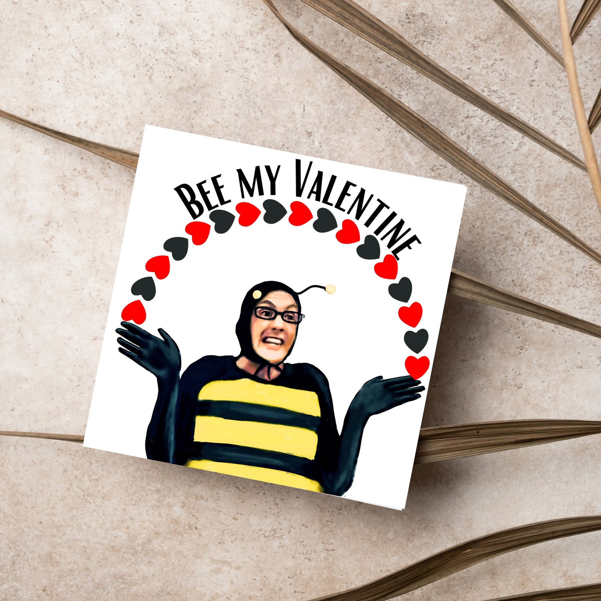 Square greetings card featuring Dean Pelton from the TV show Community, in one of his eccentric costumes dressed as a bee holding an arch of red and black hearts above his head with the text 'Bee my valentine'