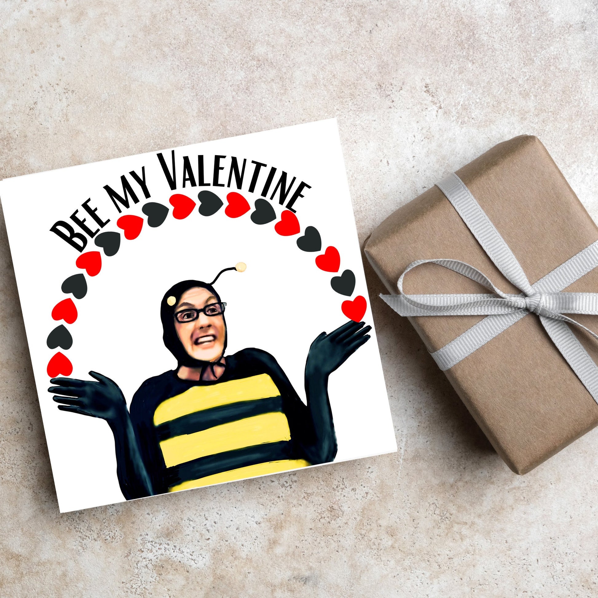 Square greetings card featuring Dean Pelton from the TV show Community, in one of his eccentric costumes dressed as a bee holding an arch of red and black hearts above his head with the text 'Bee my valentine'