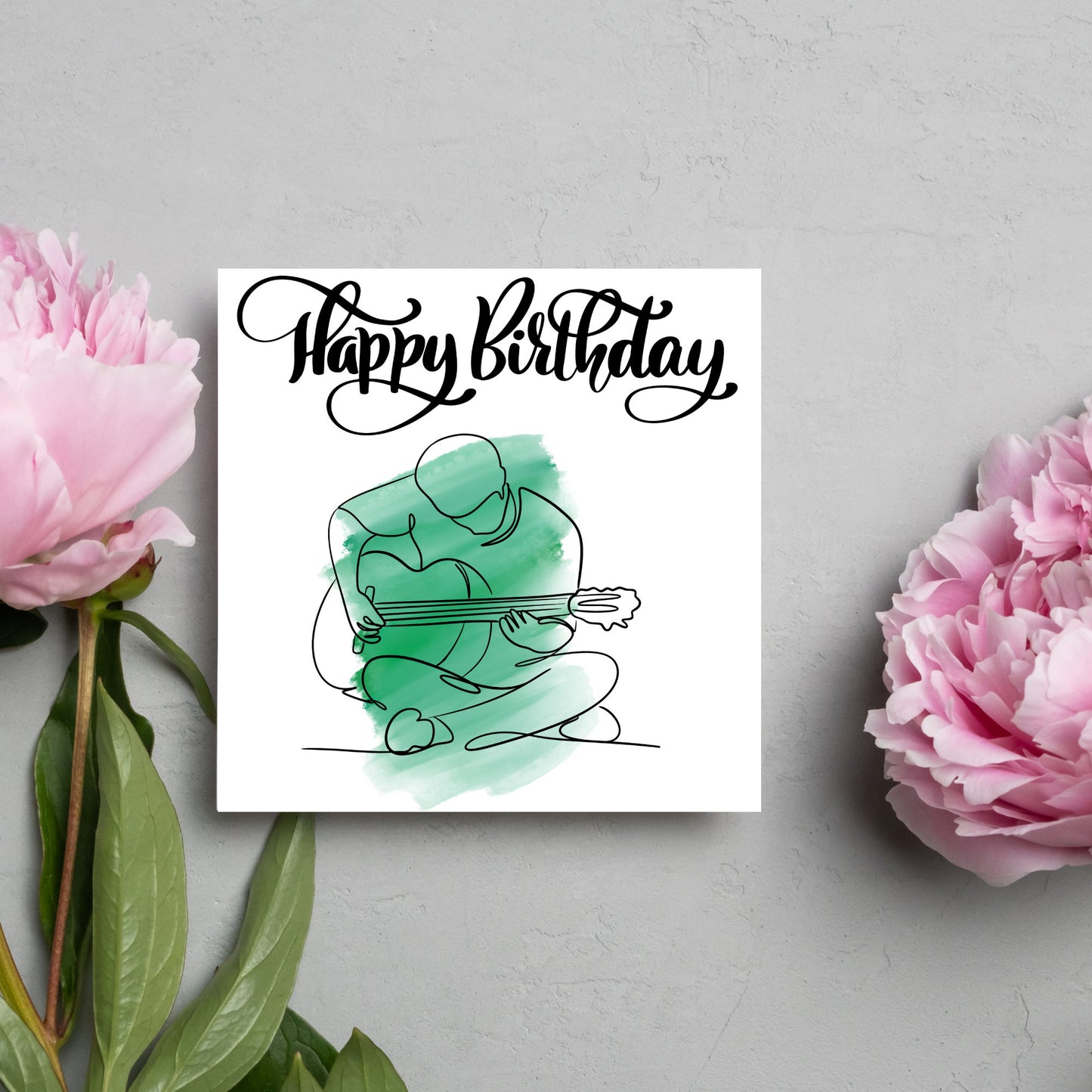 Square birthday card featuring a line drawing of a person sitting cross legged playing a guitar with a green watercolour style backdrop. The top of the card reads Happy Birthday in black swirly writing