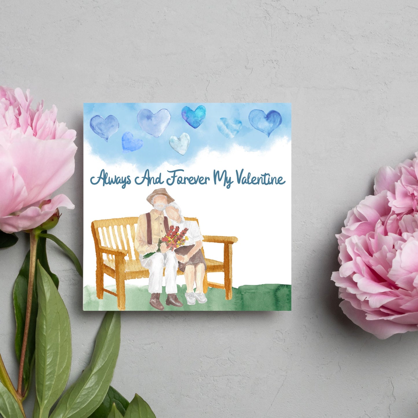 This beautiful Card has a watercolour style print with a couple sitting on a bench with flowers. Above them the blue sky has a series of cute watercolour hearts. This Cute card is perfect for valentines day for your wife or husband