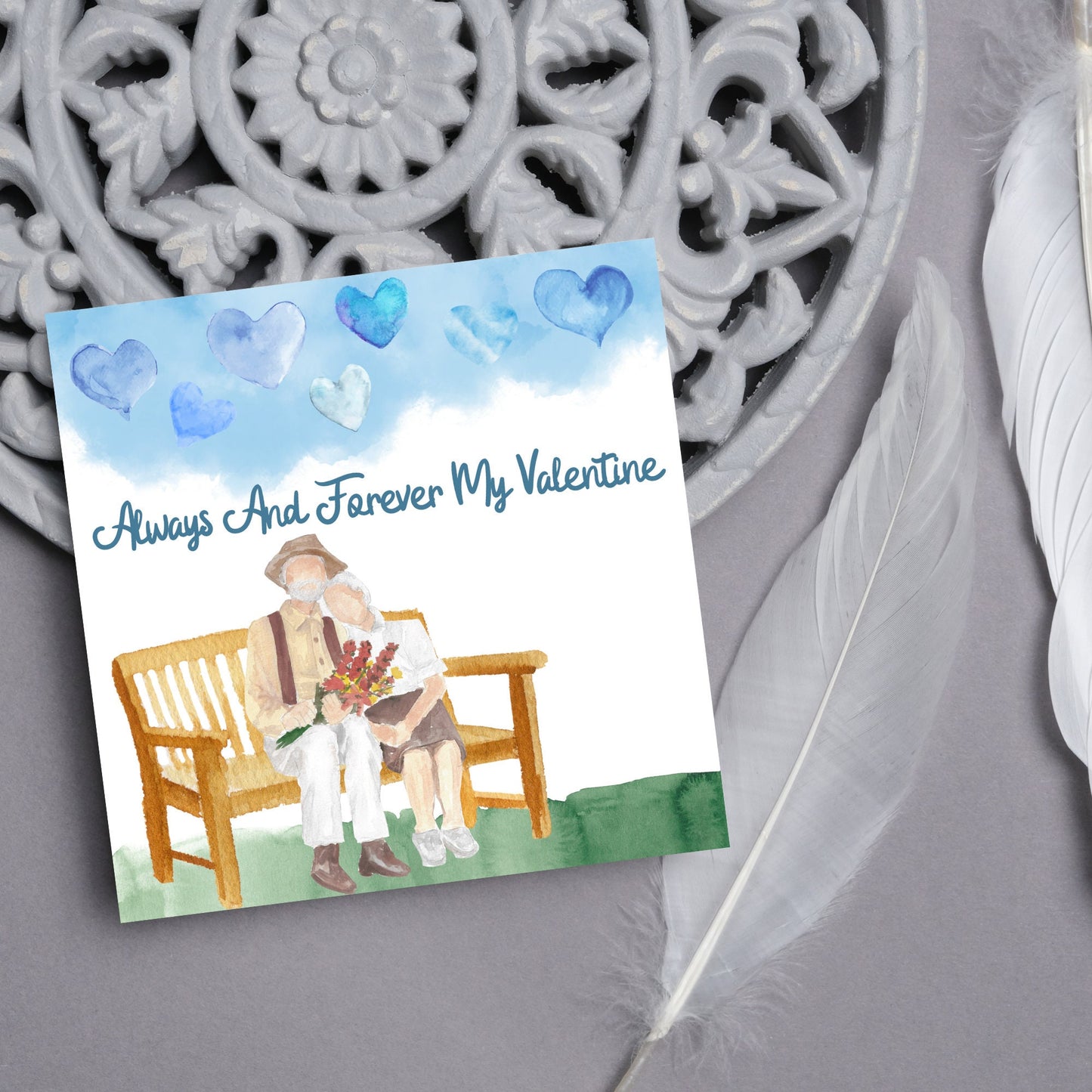This beautiful Card has a watercolour style print with a couple sitting on a bench with flowers. Above them the blue sky has a series of cute watercolour hearts. This Cute card is perfect for valentines day for your wife or husband