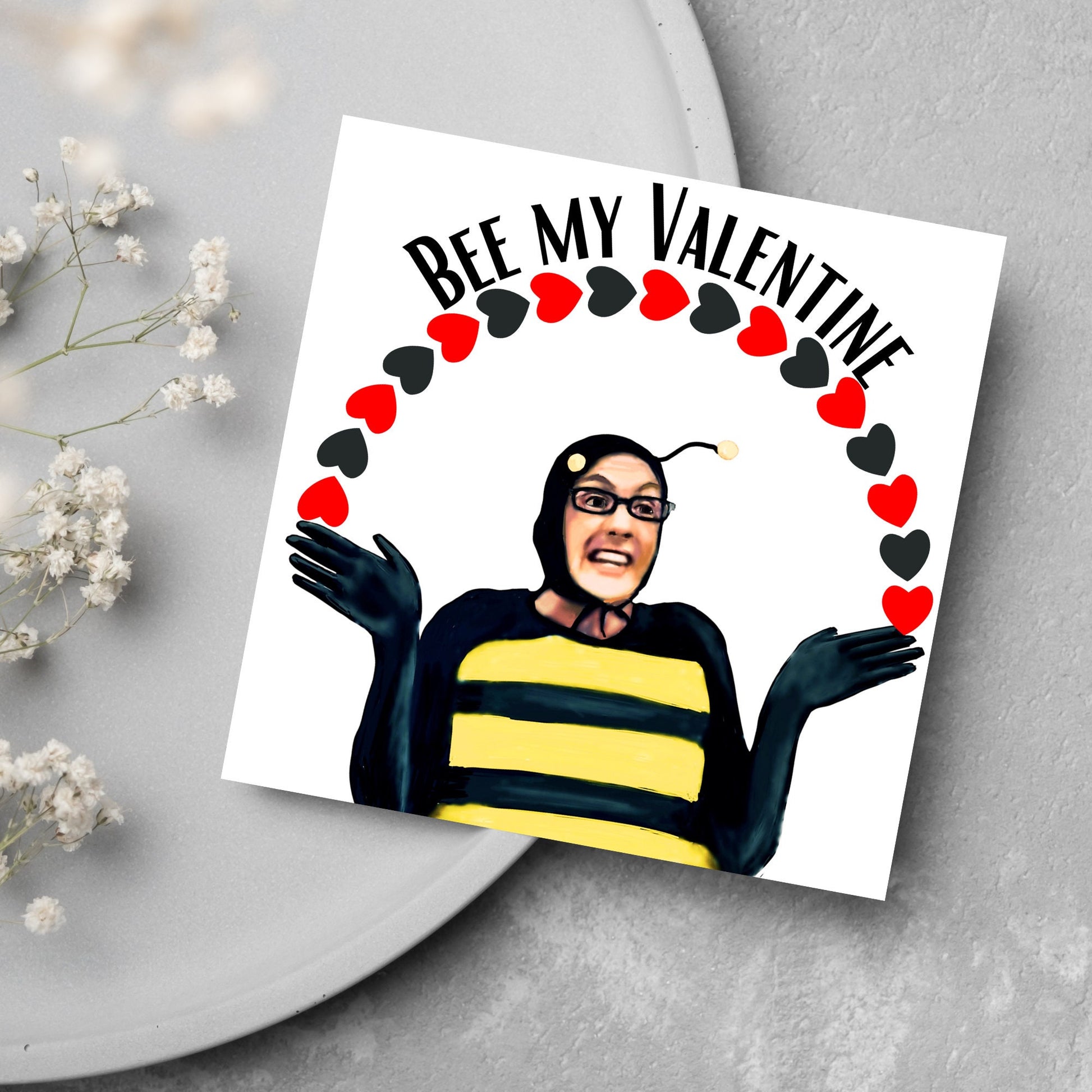 Square greetings card featuring Dean Pelton from the TV show Community, in one of his eccentric costumes dressed as a bee holding an arch of red and black hearts above his head with the text 'Bee my valentine'
