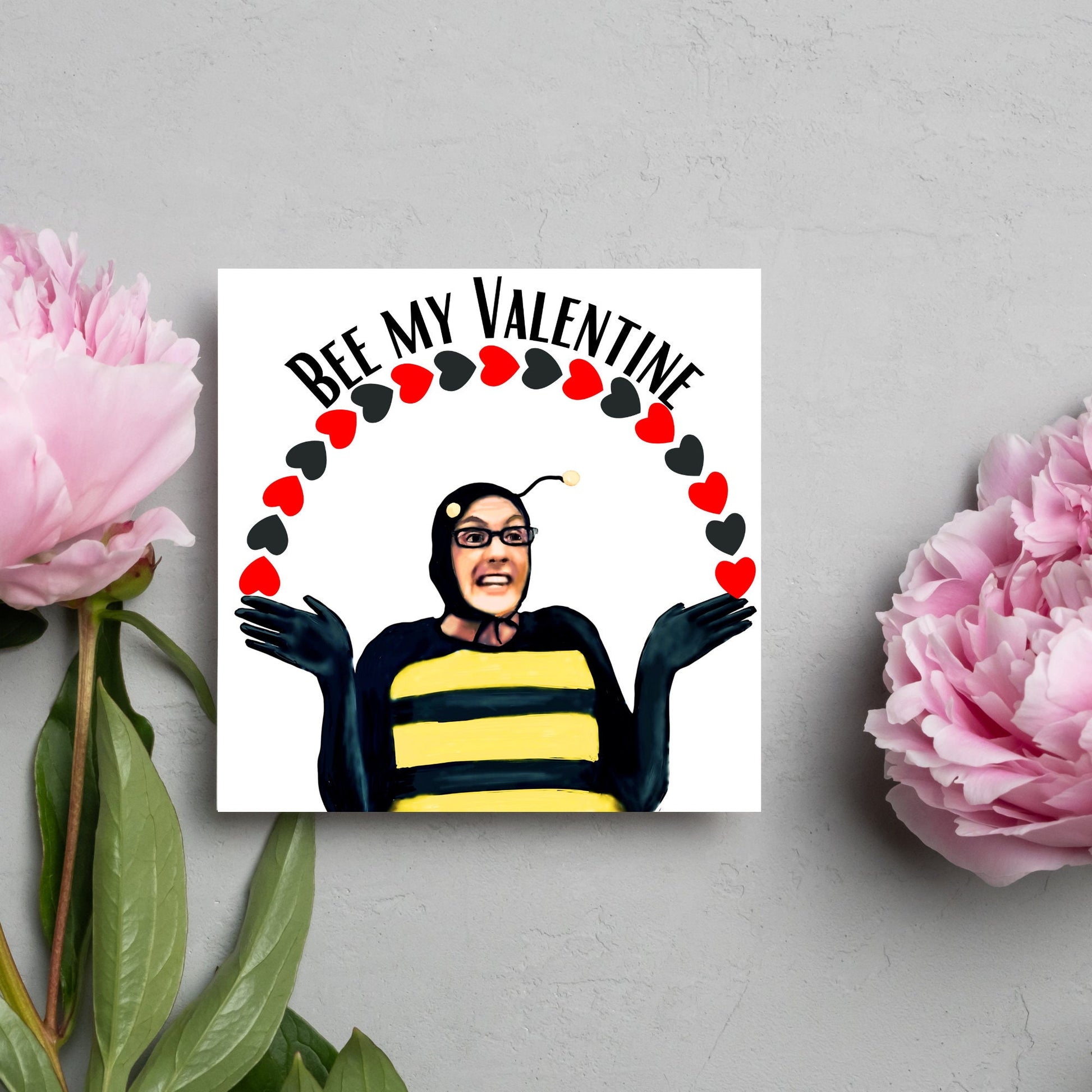 Square greetings card featuring Dean Pelton from the TV show Community, in one of his eccentric costumes dressed as a bee holding an arch of red and black hearts above his head with the text 'Bee my valentine'