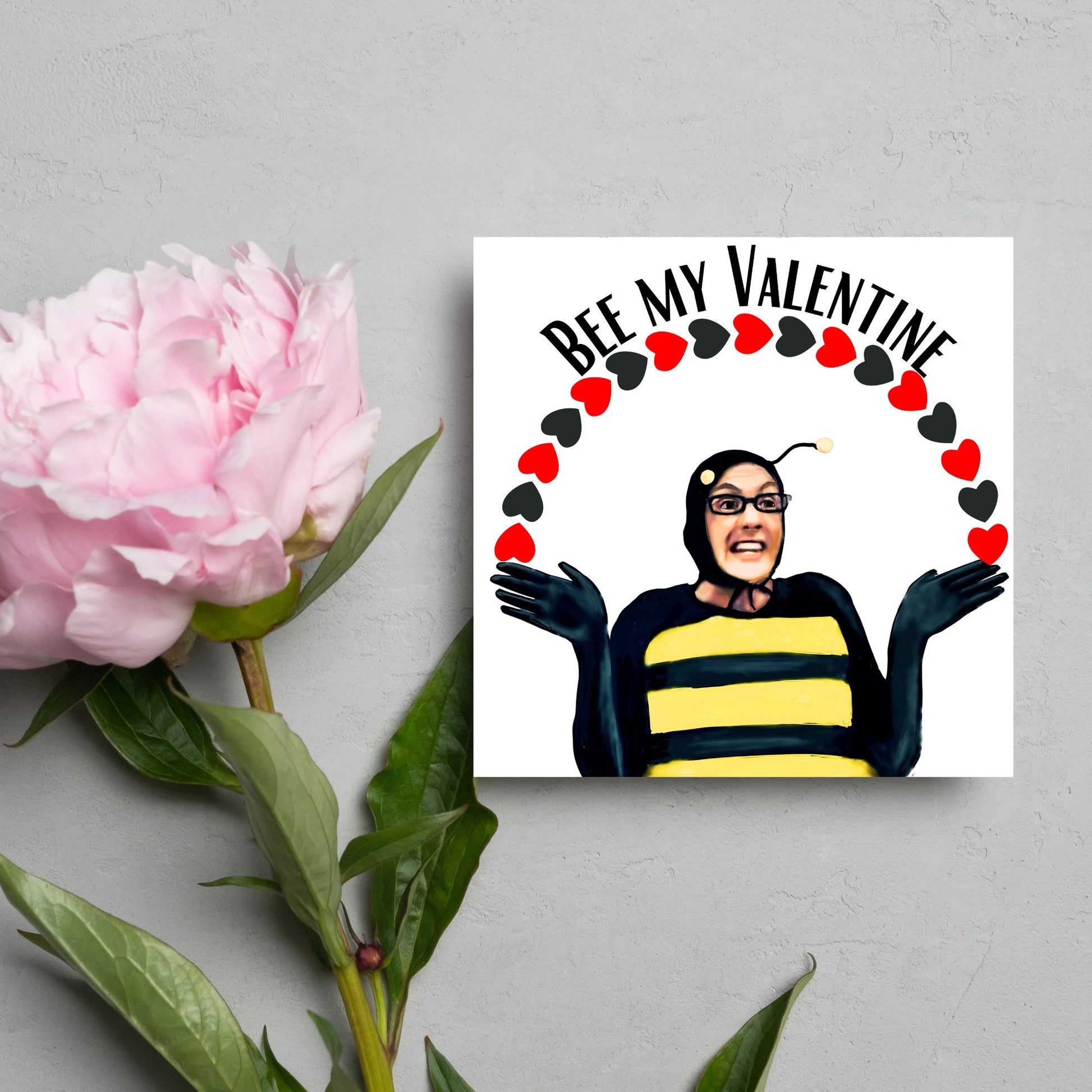 Square greetings card featuring Dean Pelton from the TV show Community, in one of his eccentric costumes dressed as a bee holding an arch of red and black hearts above his head with the text 'Bee my valentine'