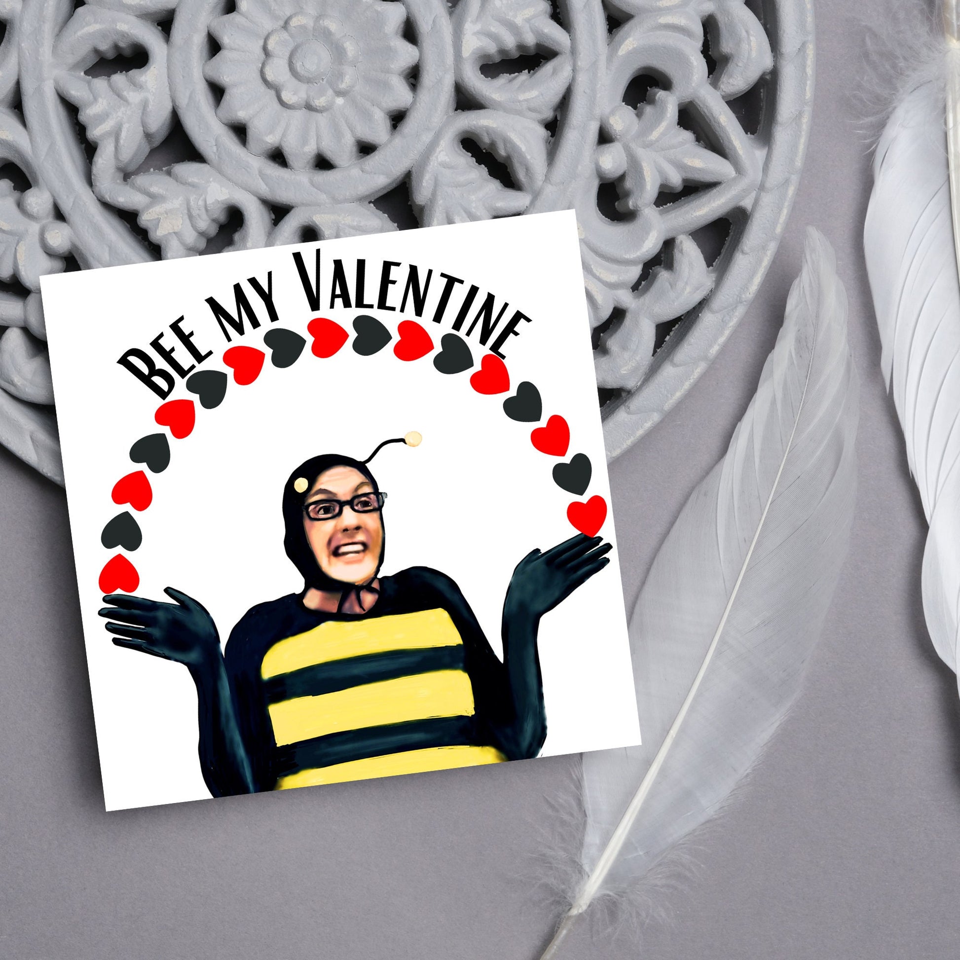 Square greetings card featuring Dean Pelton from the TV show Community, in one of his eccentric costumes dressed as a bee holding an arch of red and black hearts above his head with the text 'Bee my valentine'