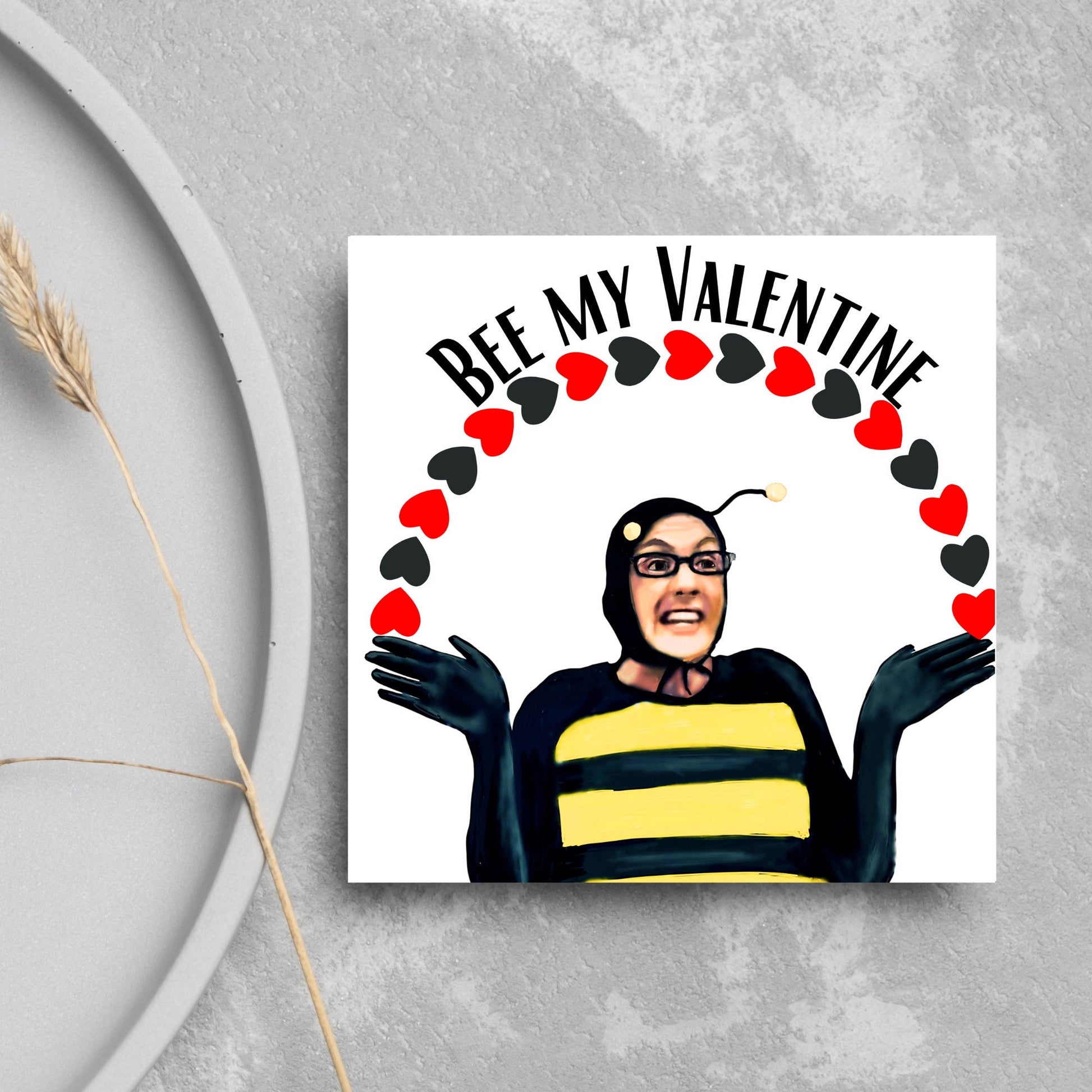 Square greetings card featuring Dean Pelton from the TV show Community, in one of his eccentric costumes dressed as a bee holding an arch of red and black hearts above his head with the text 'Bee my valentine'