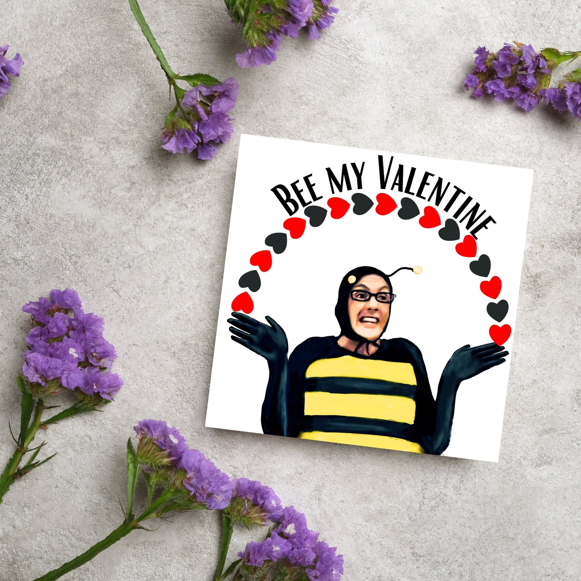Square greetings card featuring Dean Pelton from the TV show Community, in one of his eccentric costumes dressed as a bee holding an arch of red and black hearts above his head with the text 'Bee my valentine'