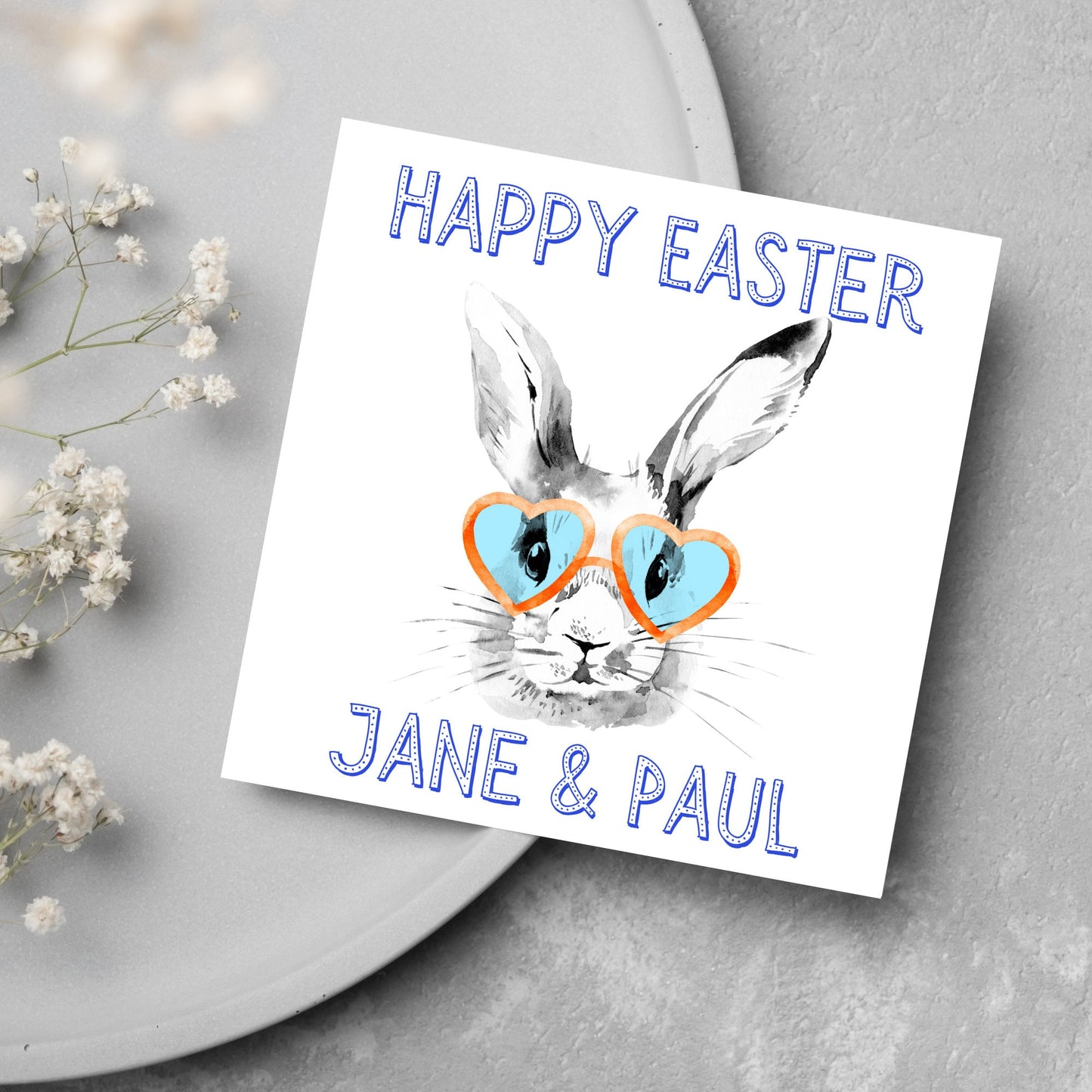 Square card with a simple watercolour style illustration of a white rabbit wearing orange and blue heart shaped sunglasses. The text reads 'Happy Easter Jane and Paul'