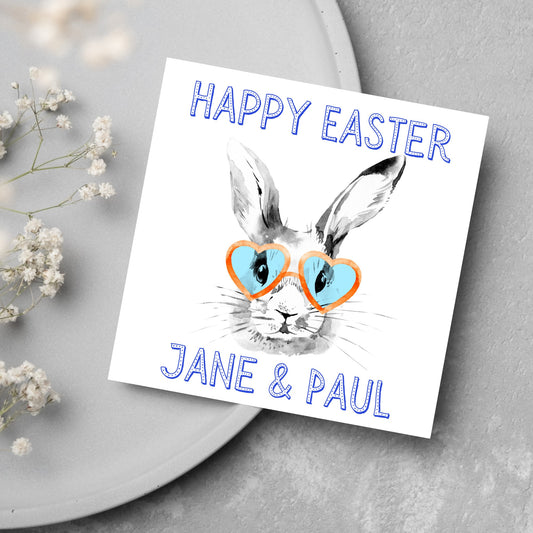 Square card with a simple watercolour style illustration of a white rabbit wearing orange and blue heart shaped sunglasses. The text reads 'Happy Easter Jane and Paul'