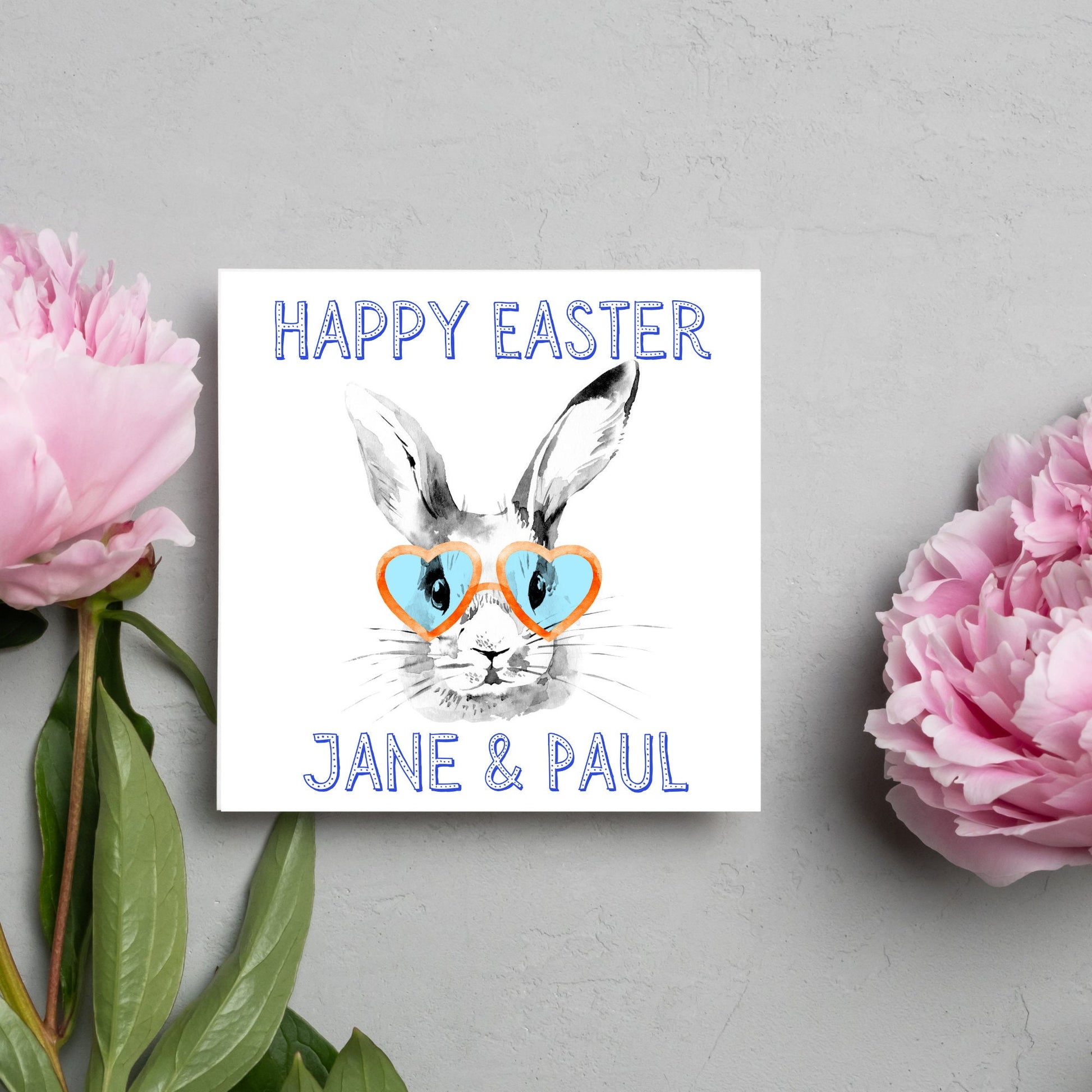 Square card with a simple watercolour style illustration of a white rabbit wearing orange and blue heart shaped sunglasses. The text reads 'Happy Easter Jane and Paul'
