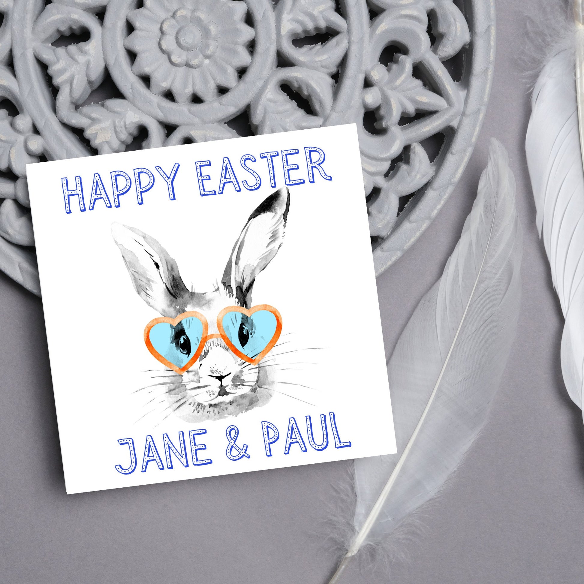 Square card with a simple watercolour style illustration of a white rabbit wearing orange and blue heart shaped sunglasses. The text reads 'Happy Easter Jane and Paul'