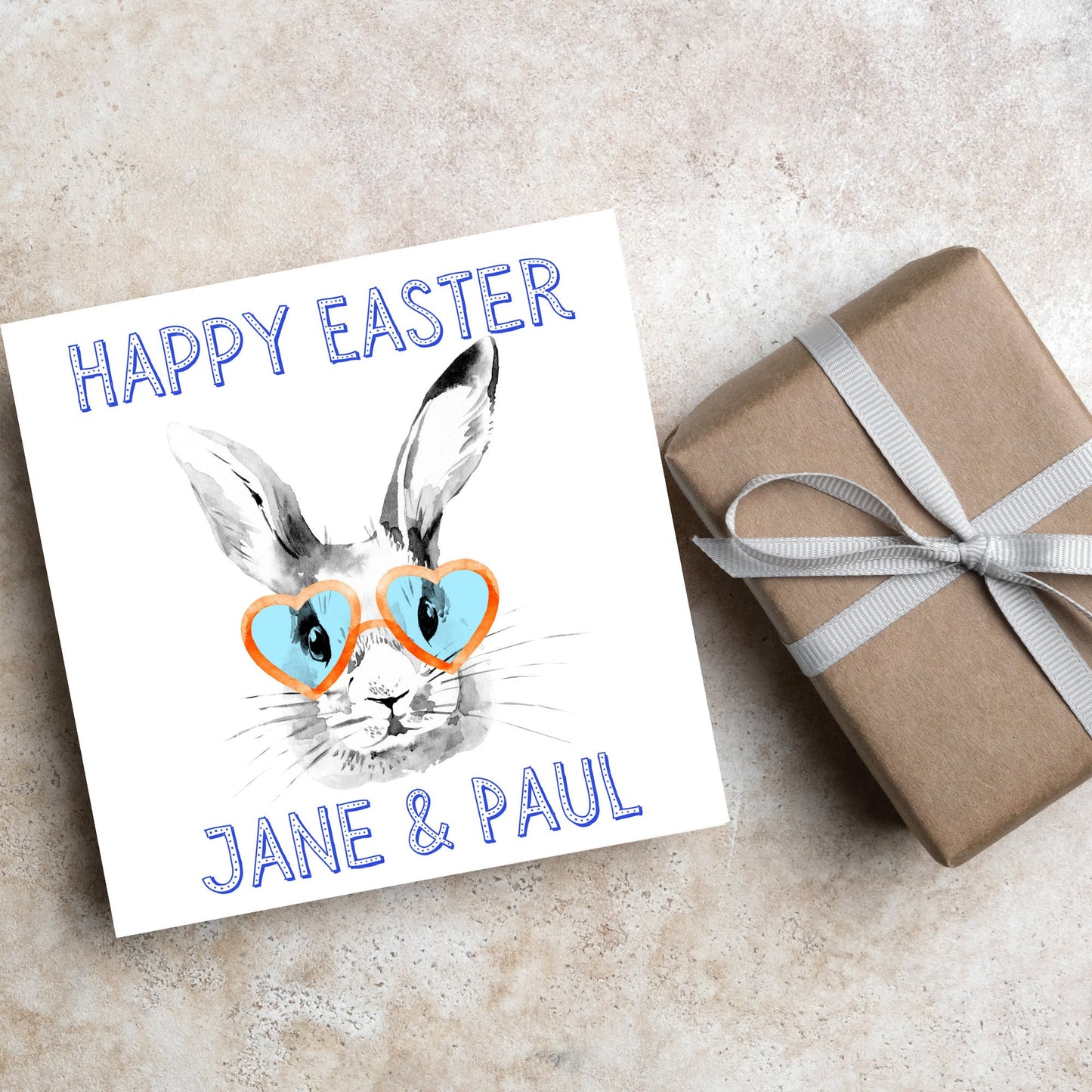 Square card with a simple watercolour style illustration of a white rabbit wearing orange and blue heart shaped sunglasses. The text reads 'Happy Easter Jane and Paul'