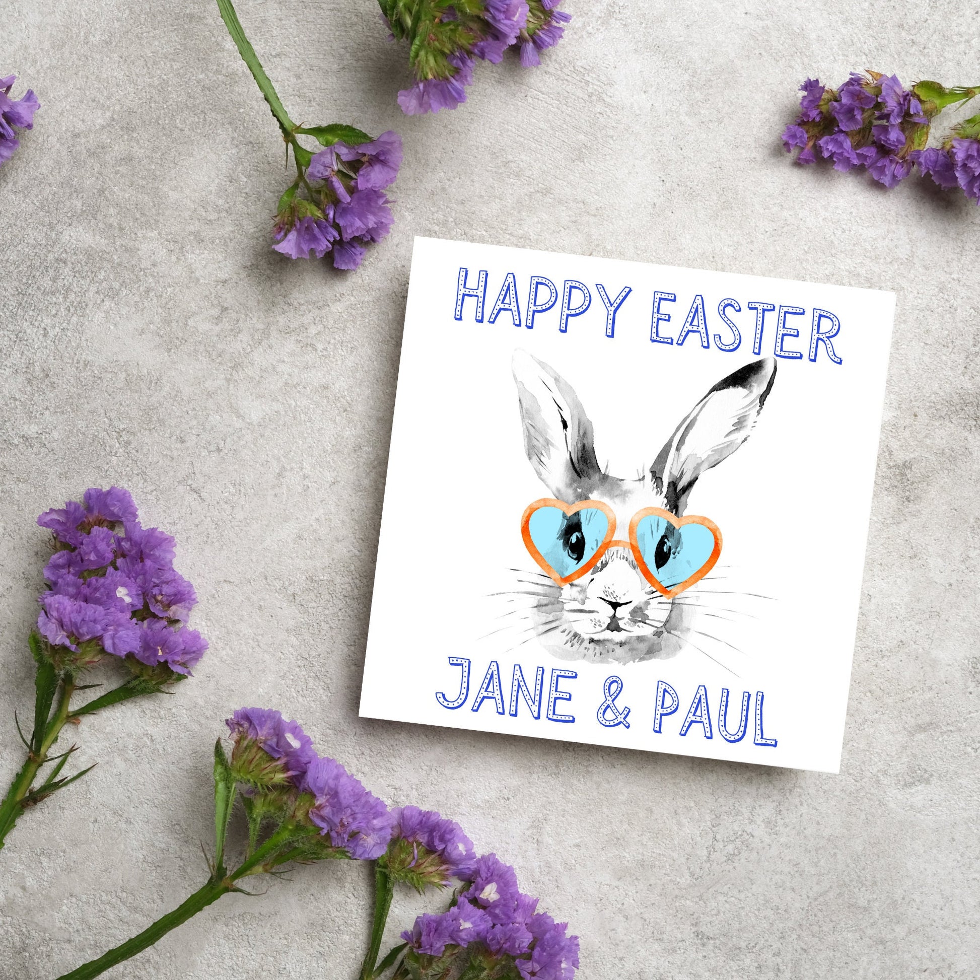 Square card with a simple watercolour style illustration of a white rabbit wearing orange and blue heart shaped sunglasses. The text reads 'Happy Easter Jane and Paul'