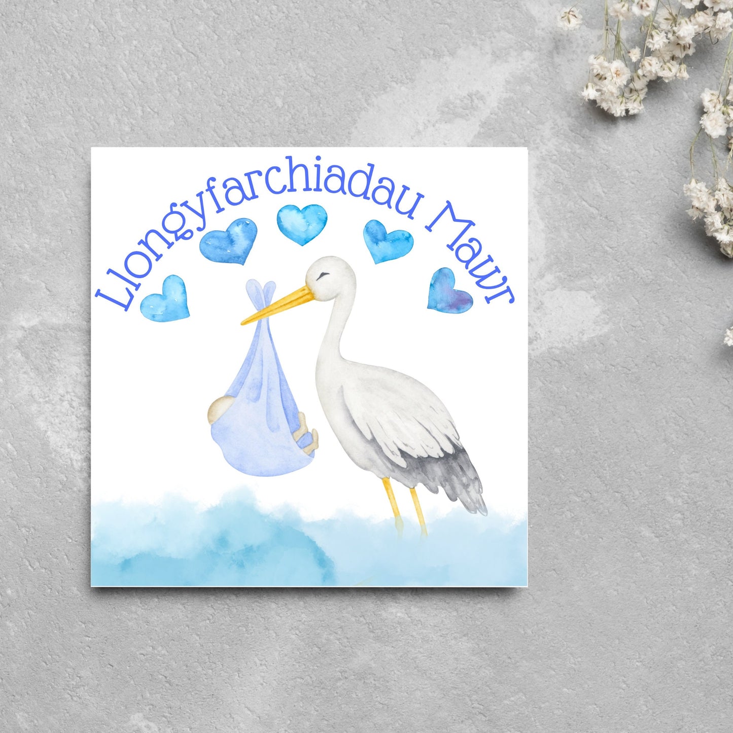 Image of a square greetings card with a stork delivering a baby in a blue blanket. THere are 5 blue watercolour hearts above the stork and the at the top is blue text 'llongyfarchiadau Mawr' which translates from welsh as many congratulations