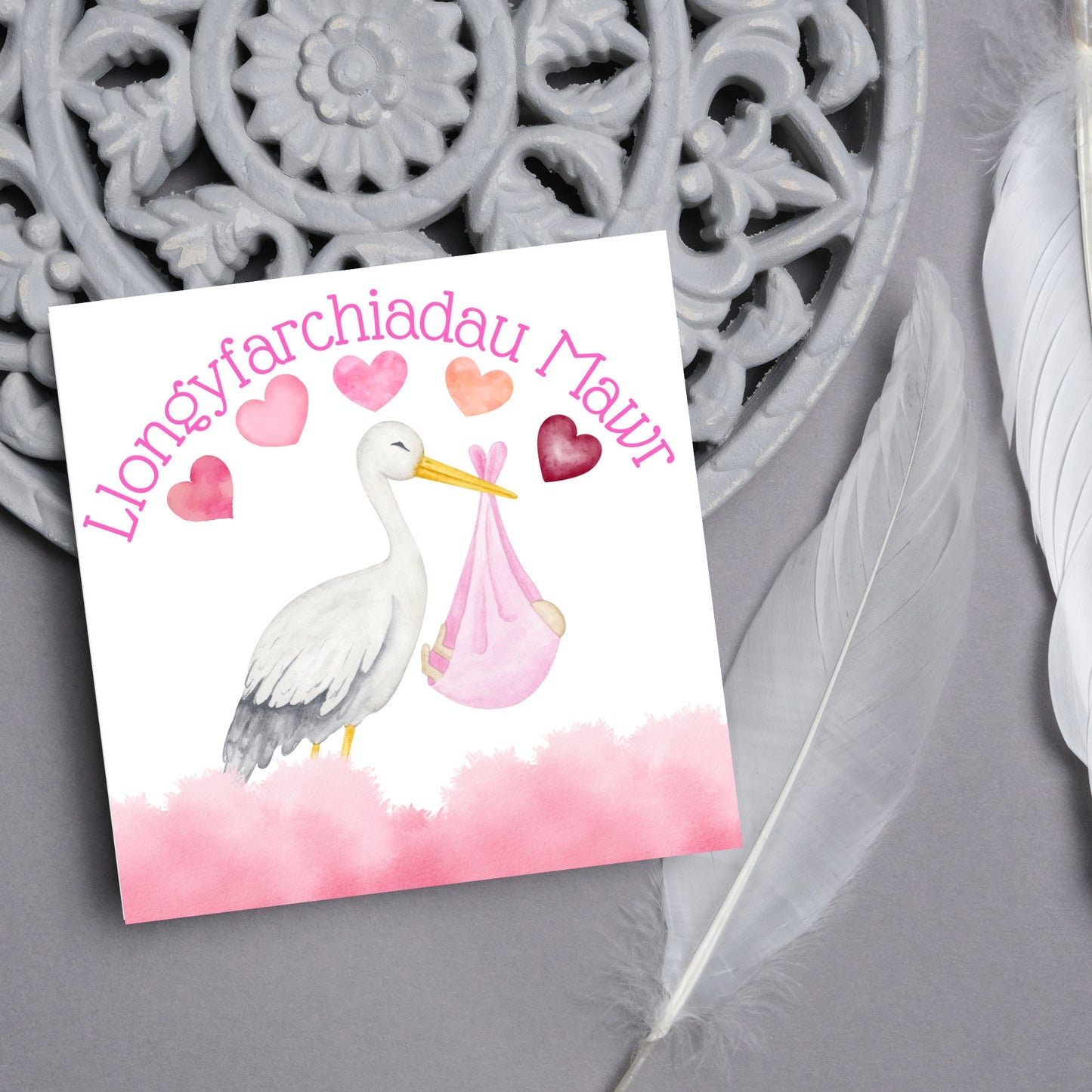 Image of a square greetings card with a stork delivering a baby in a pink blanket. There are 5 pink watercolour hearts above the stork and the at the top is pink text 'llongyfarchiadau Mawr' which translates from welsh as many congratulations