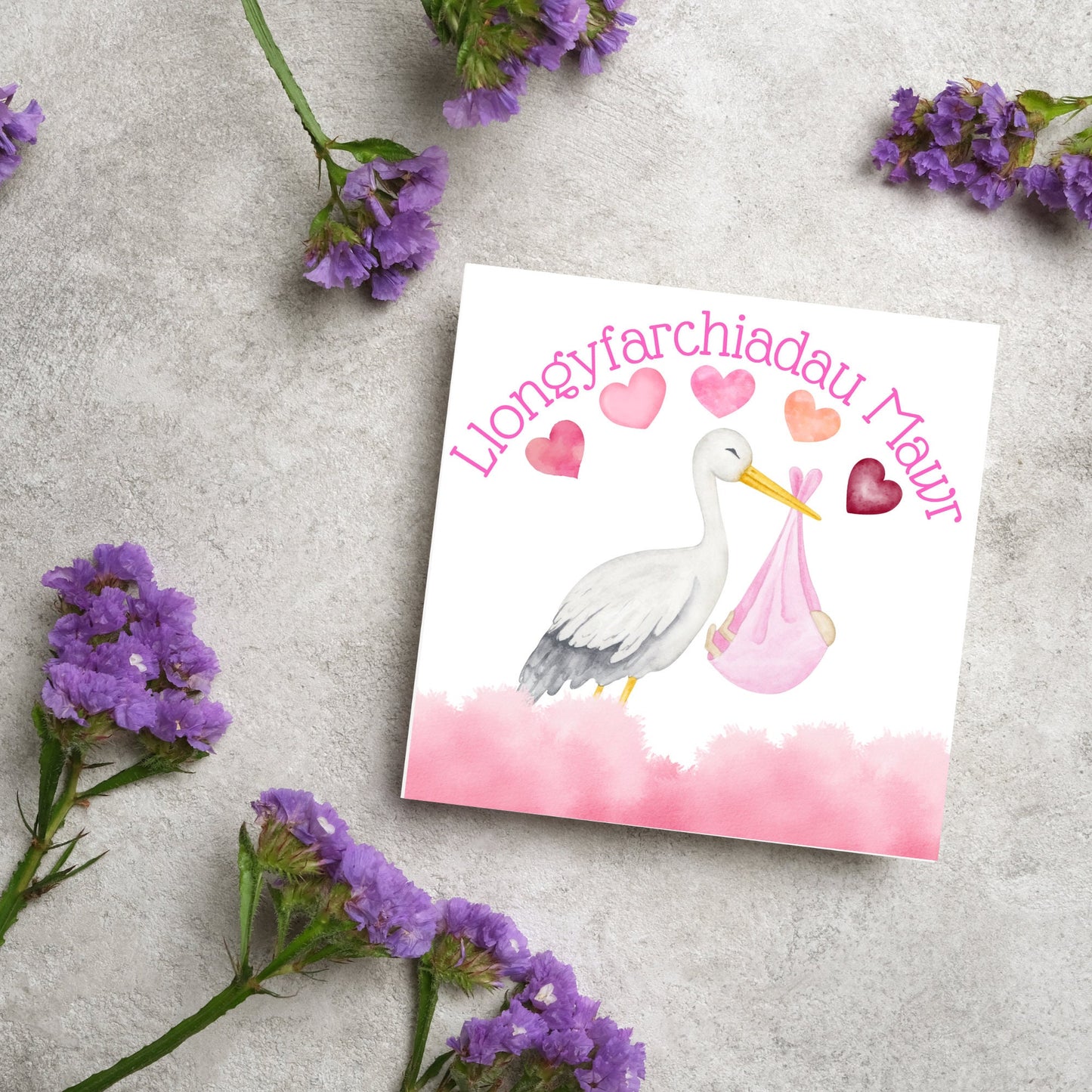 Image of a square greetings card with a stork delivering a baby in a pink blanket. There are 5 pink watercolour hearts above the stork and the at the top is pink text 'llongyfarchiadau Mawr' which translates from welsh as many congratulations