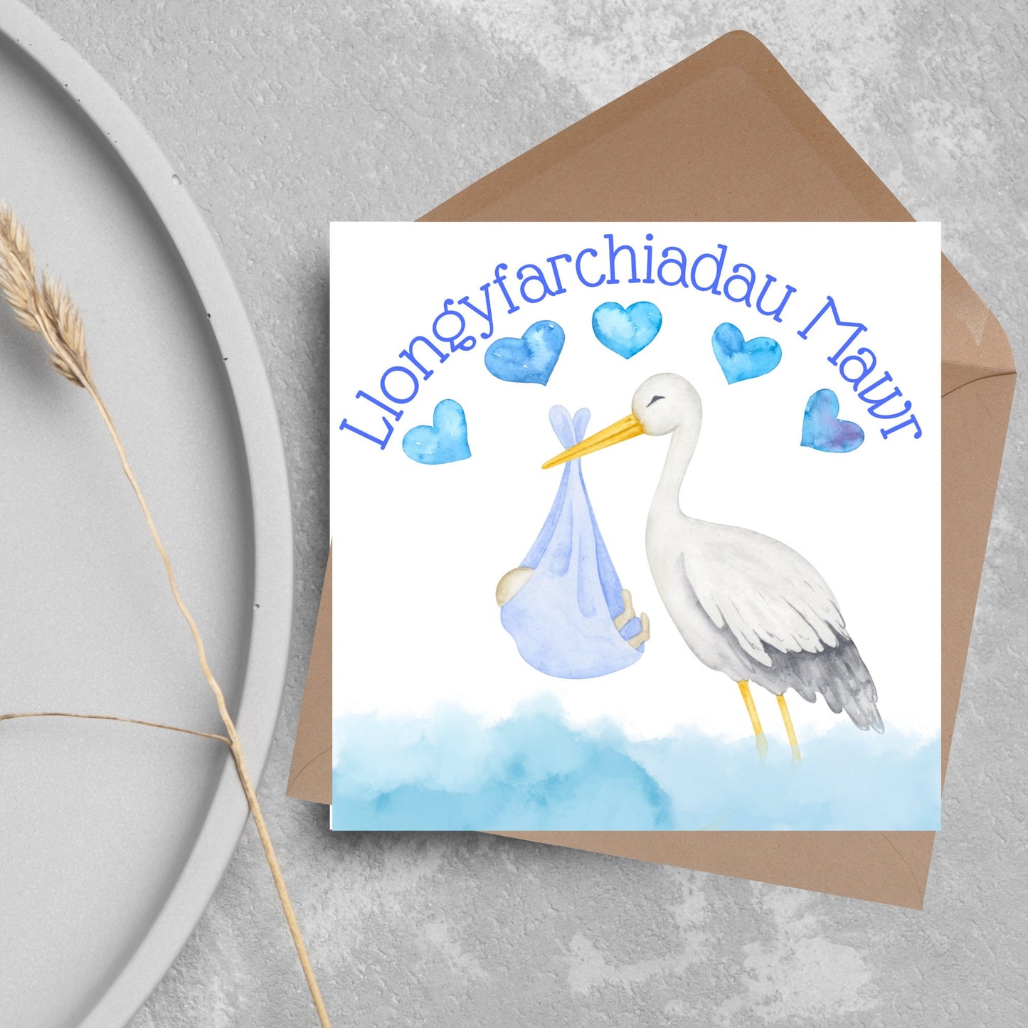 Image of a square greetings card with a stork delivering a baby in a blue blanket. THere are 5 blue watercolour hearts above the stork and the at the top is blue text 'llongyfarchiadau Mawr' which translates from welsh as many congratulations
