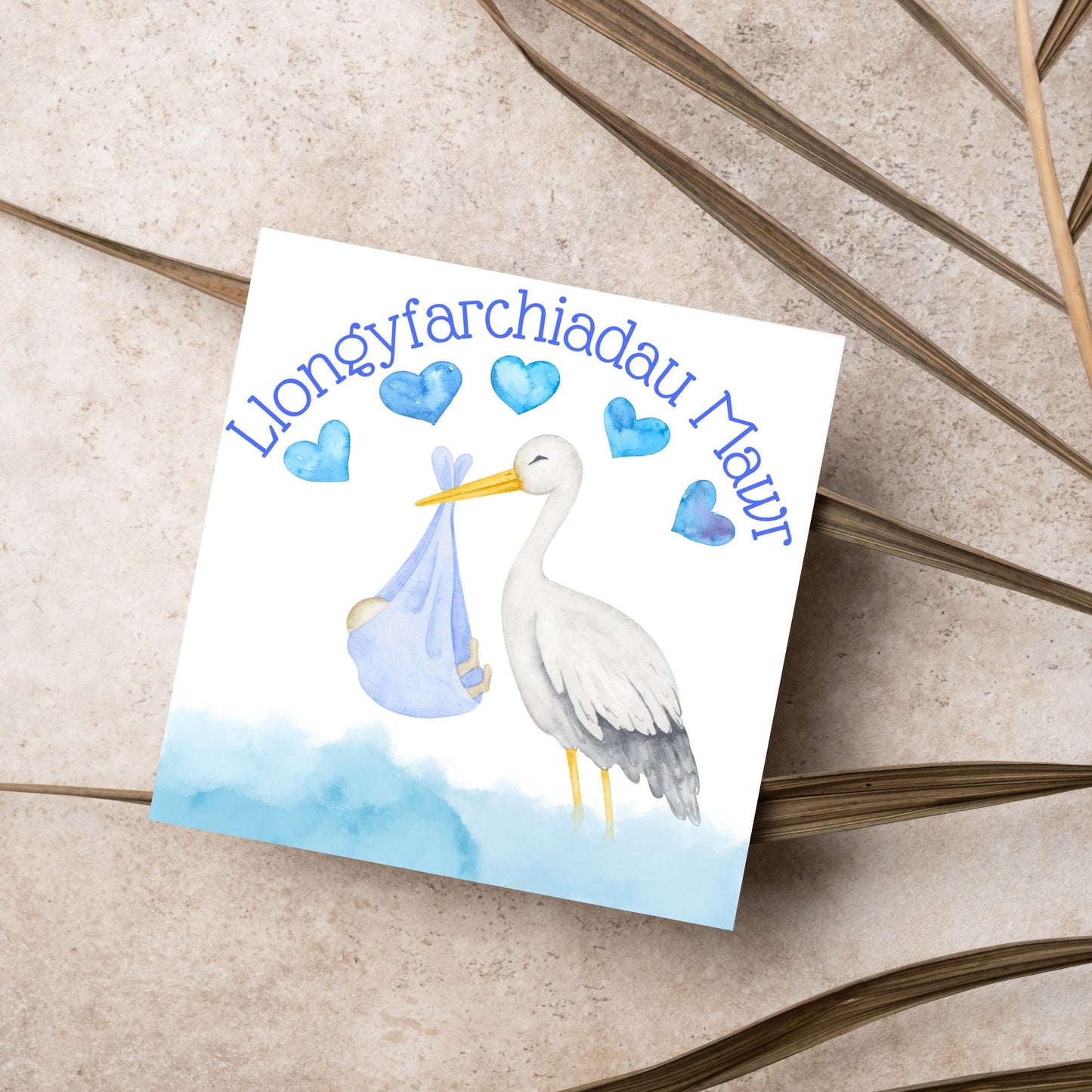 Image of a square greetings card with a stork delivering a baby in a blue blanket. THere are 5 blue watercolour hearts above the stork and the at the top is blue text 'llongyfarchiadau Mawr' which translates from welsh as many congratulations