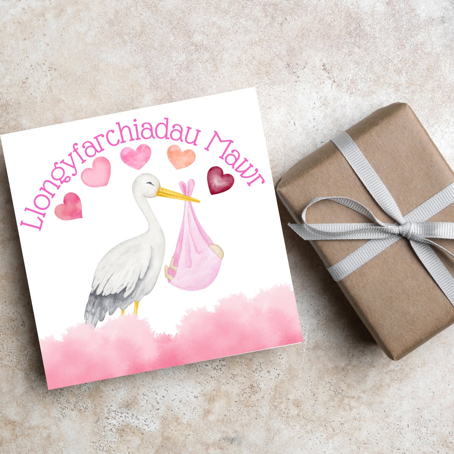 Image of a square greetings card with a stork delivering a baby in a pink blanket. There are 5 pink watercolour hearts above the stork and the at the top is pink text 'llongyfarchiadau Mawr' which translates from welsh as many congratulations