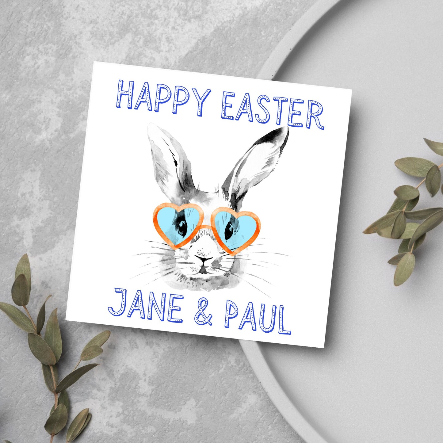 Square card with a simple watercolour style illustration of a white rabbit wearing orange and blue heart shaped sunglasses. The text reads 'Happy Easter Jane and Paul'