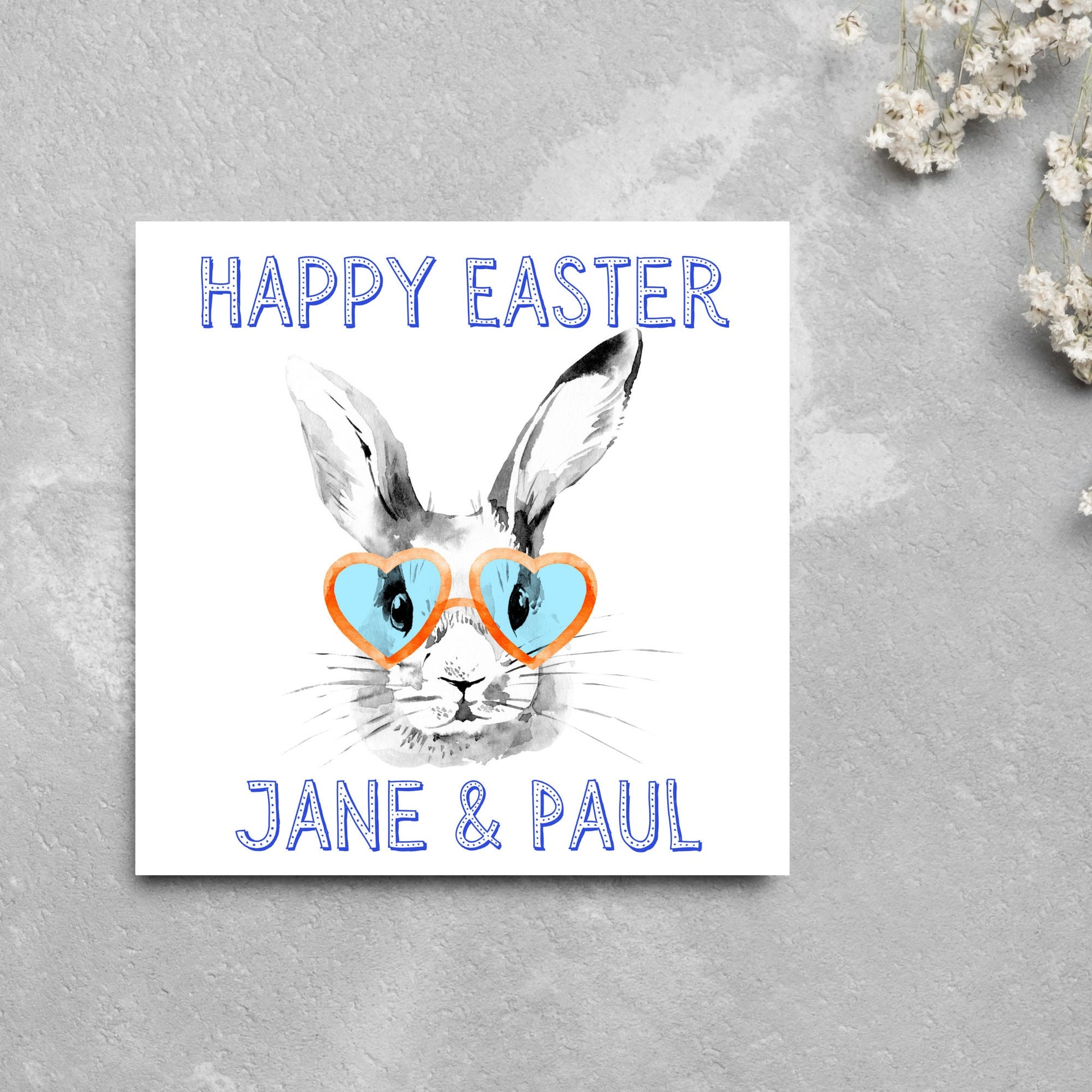 Square card with a simple watercolour style illustration of a white rabbit wearing orange and blue heart shaped sunglasses. The text reads 'Happy Easter Jane and Paul'