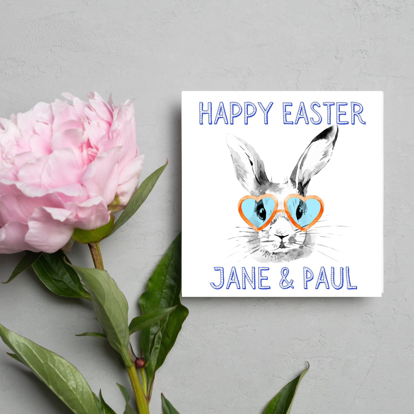 Square card with a simple watercolour style illustration of a white rabbit wearing orange and blue heart shaped sunglasses. The text reads 'Happy Easter Jane and Paul'