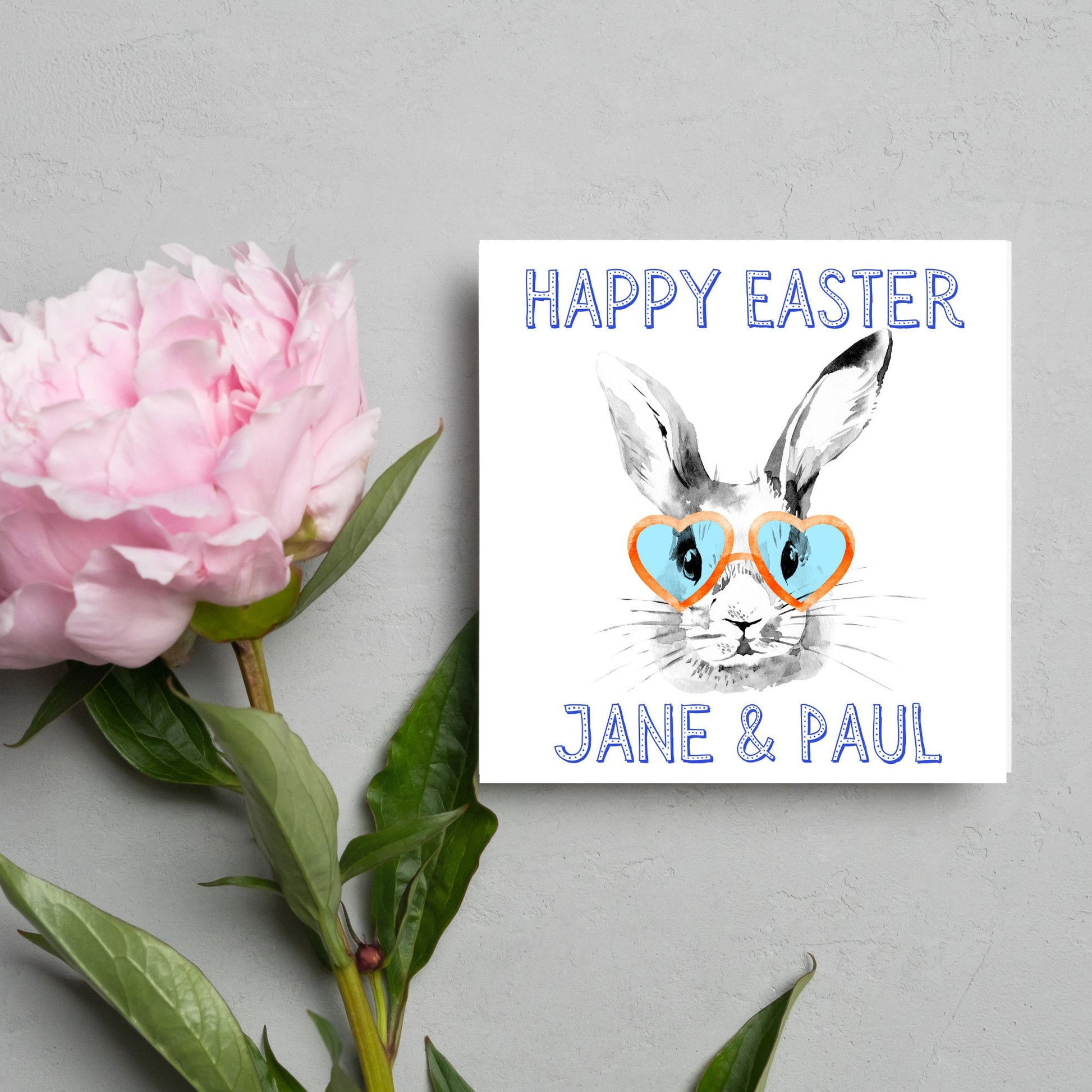Square card with a simple watercolour style illustration of a white rabbit wearing orange and blue heart shaped sunglasses. The text reads 'Happy Easter Jane and Paul'