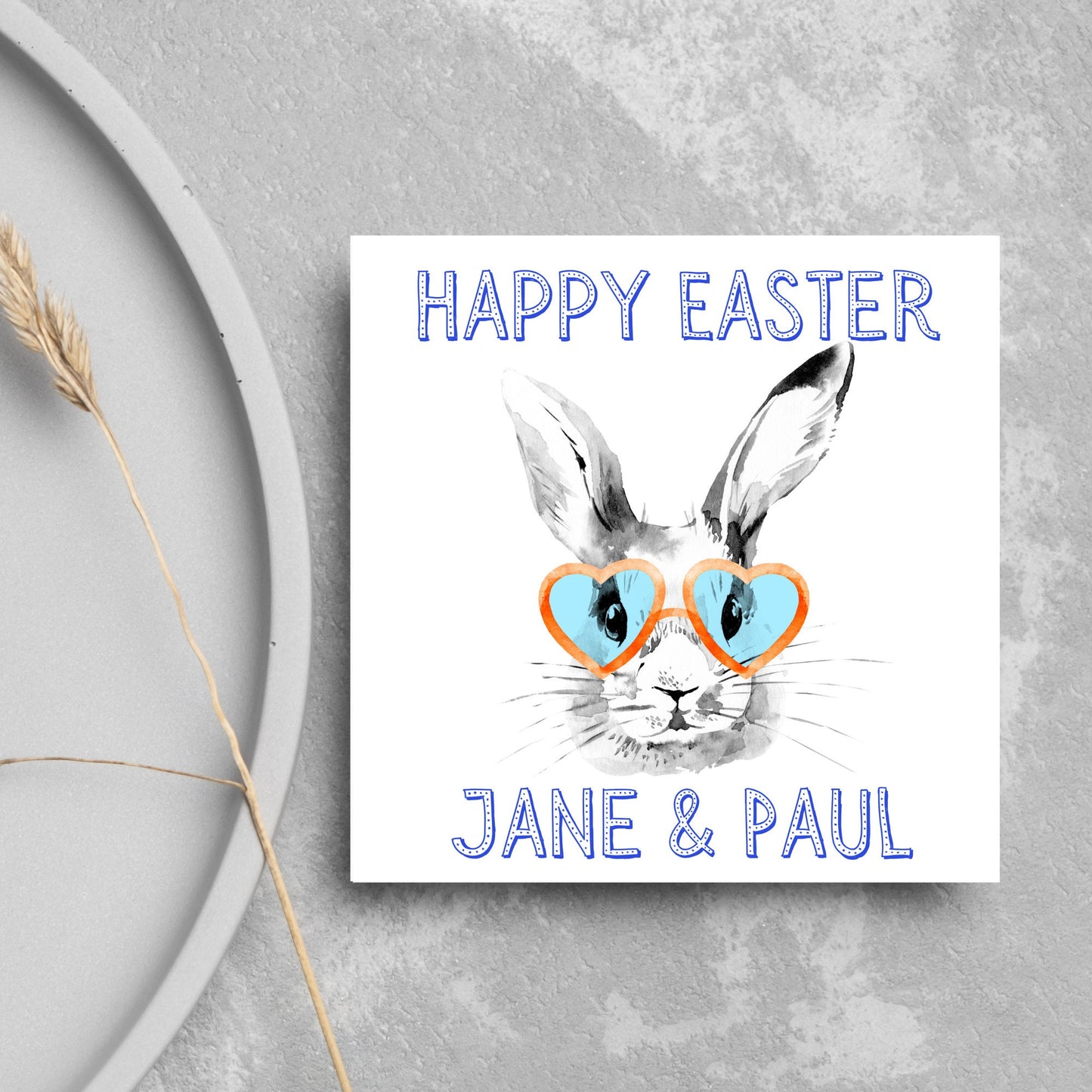 Square card with a simple watercolour style illustration of a white rabbit wearing orange and blue heart shaped sunglasses. The text reads 'Happy Easter Jane and Paul'