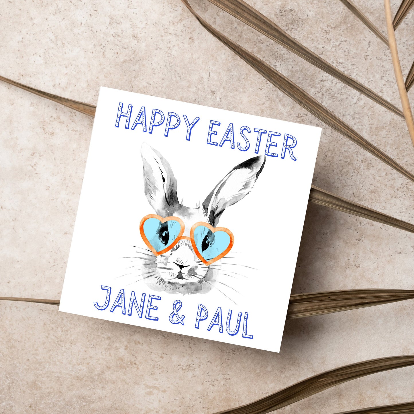 Square card with a simple watercolour style illustration of a white rabbit wearing orange and blue heart shaped sunglasses. The text reads 'Happy Easter Jane and Paul'