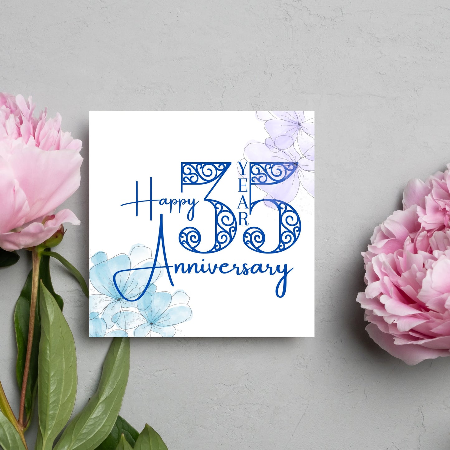 Square Card with white background, there are blue faded watercolour flowers on each corner and dark blue text reads 'Happy 35 year anniversary' the words are in a script font and the number is an ornate filigree style