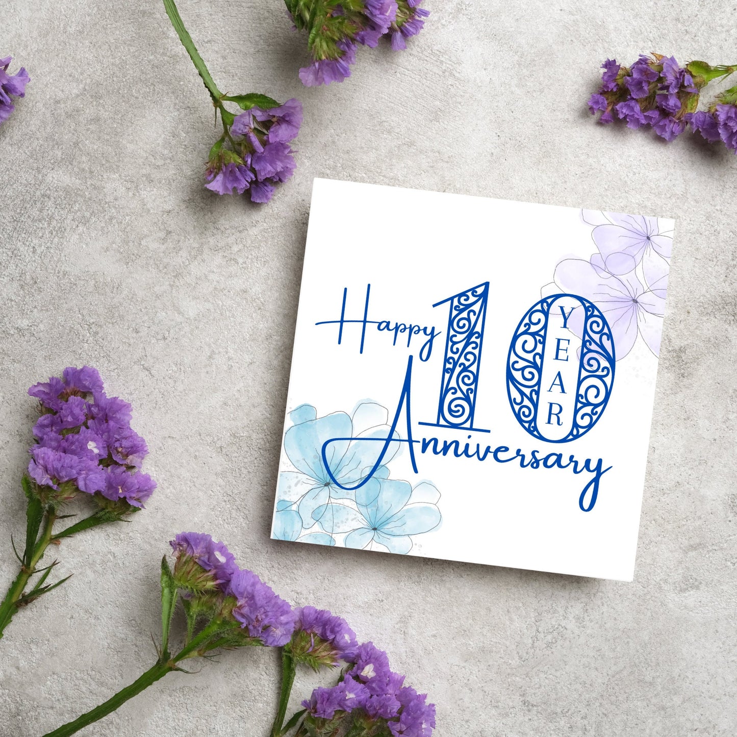 Square Card with white background, there are blue faded watercolour flowers on each corner and dark blue text reads 'Happy 10 year anniversary' the words are in a script font and the number is an ornate filigree style
