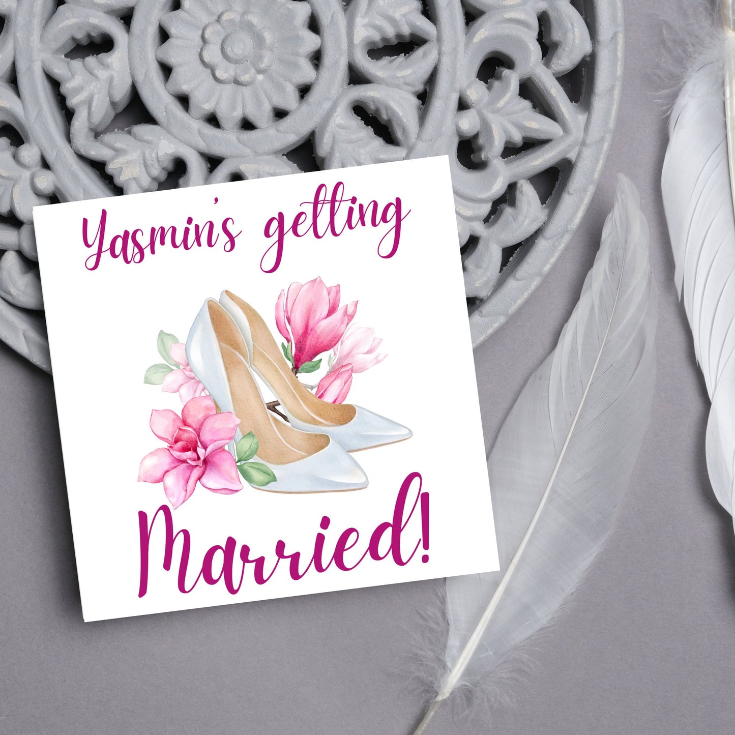 Square card with a watercolour image of white wedding shoes surrounded by pink flowers with pink text that reads Yasmin's getting married in calligraphy style font
