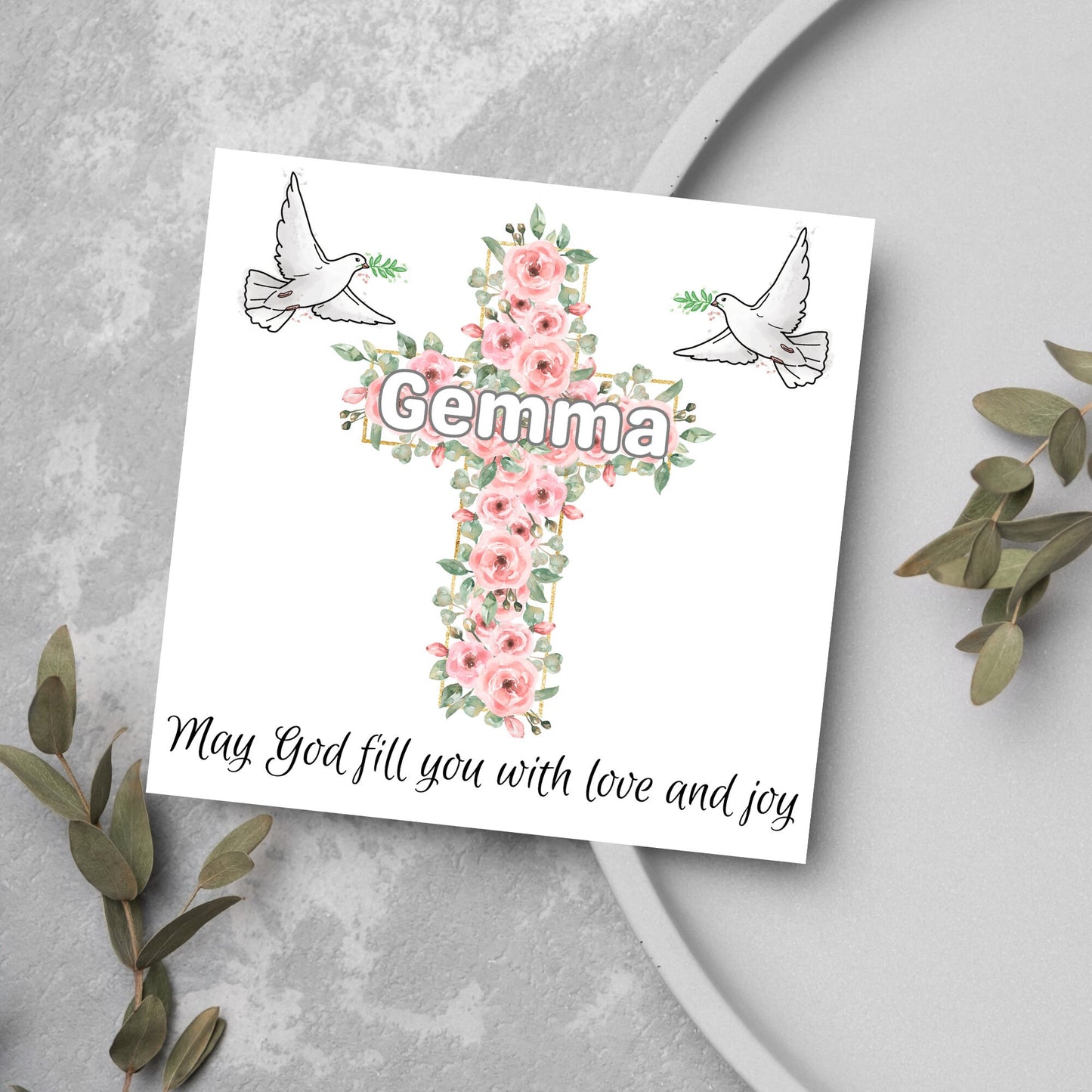 Square white card with a cross made of pink roses at the centre with the chosen name written across in white. either side of the cross is a white dove flying. At the bottom the text 'may god fill you with love and joy' is written