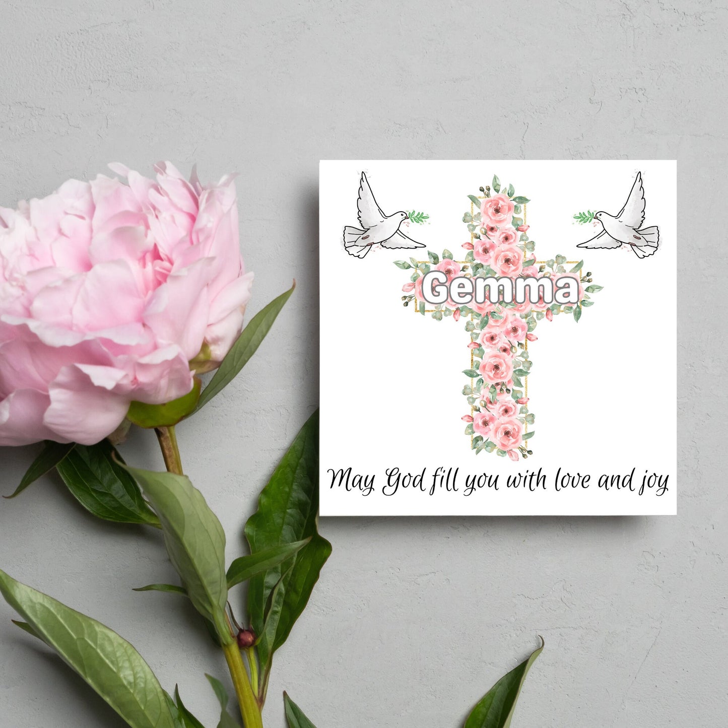Square white card with a cross made of pink roses at the centre with the chosen name written across in white. either side of the cross is a white dove flying. At the bottom the text 'may god fill you with love and joy' is written