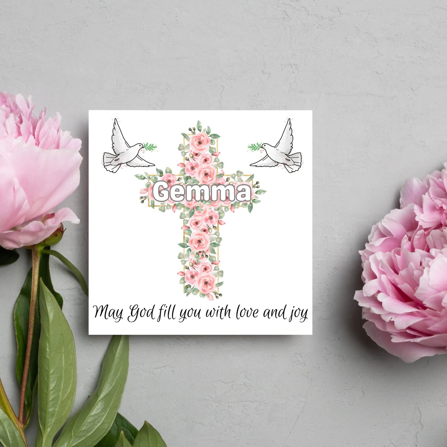 Square white card with a cross made of pink roses at the centre with the chosen name written across in white. either side of the cross is a white dove flying. At the bottom the text 'may god fill you with love and joy' is written