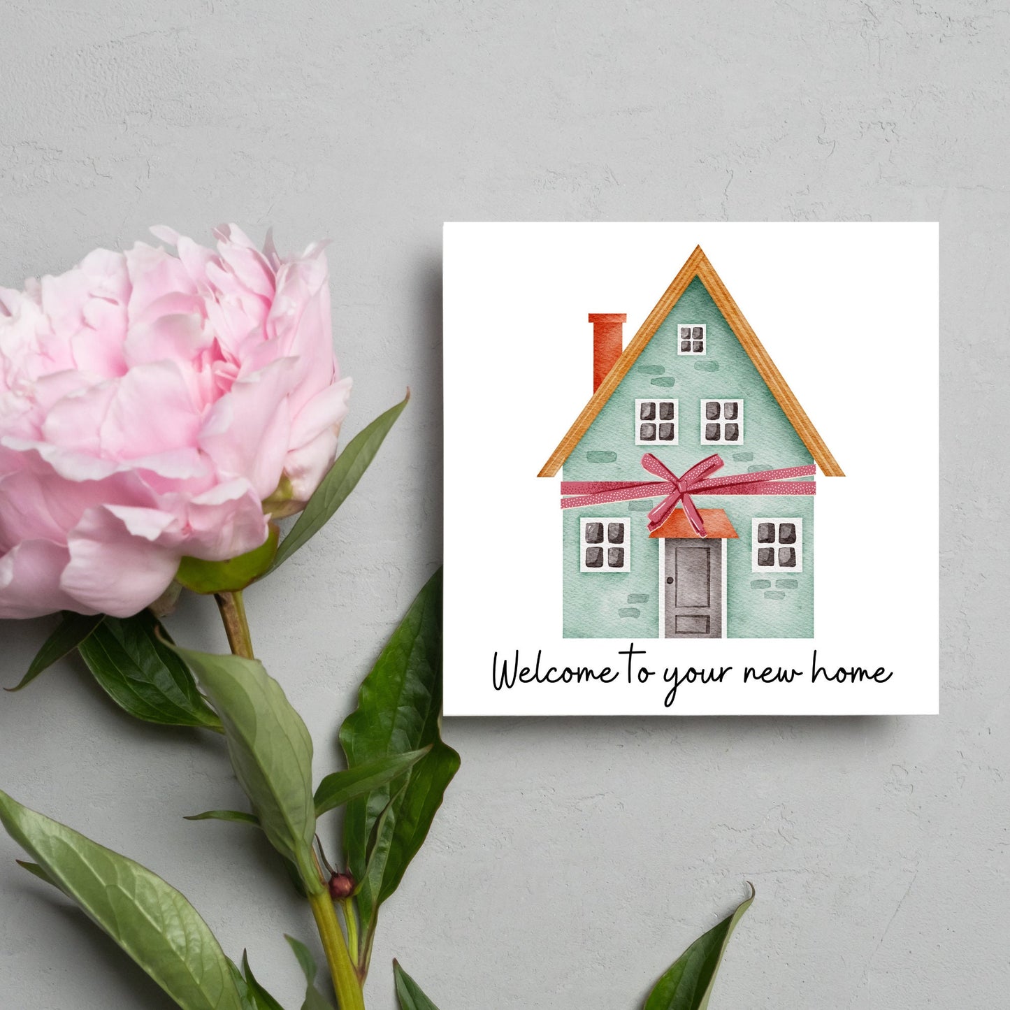 Square card with white background and a watercolour style illlustration of a green three storey house with a red bow wrapped around it. Underneath the house ina script font are the words 'welcome to your new home'