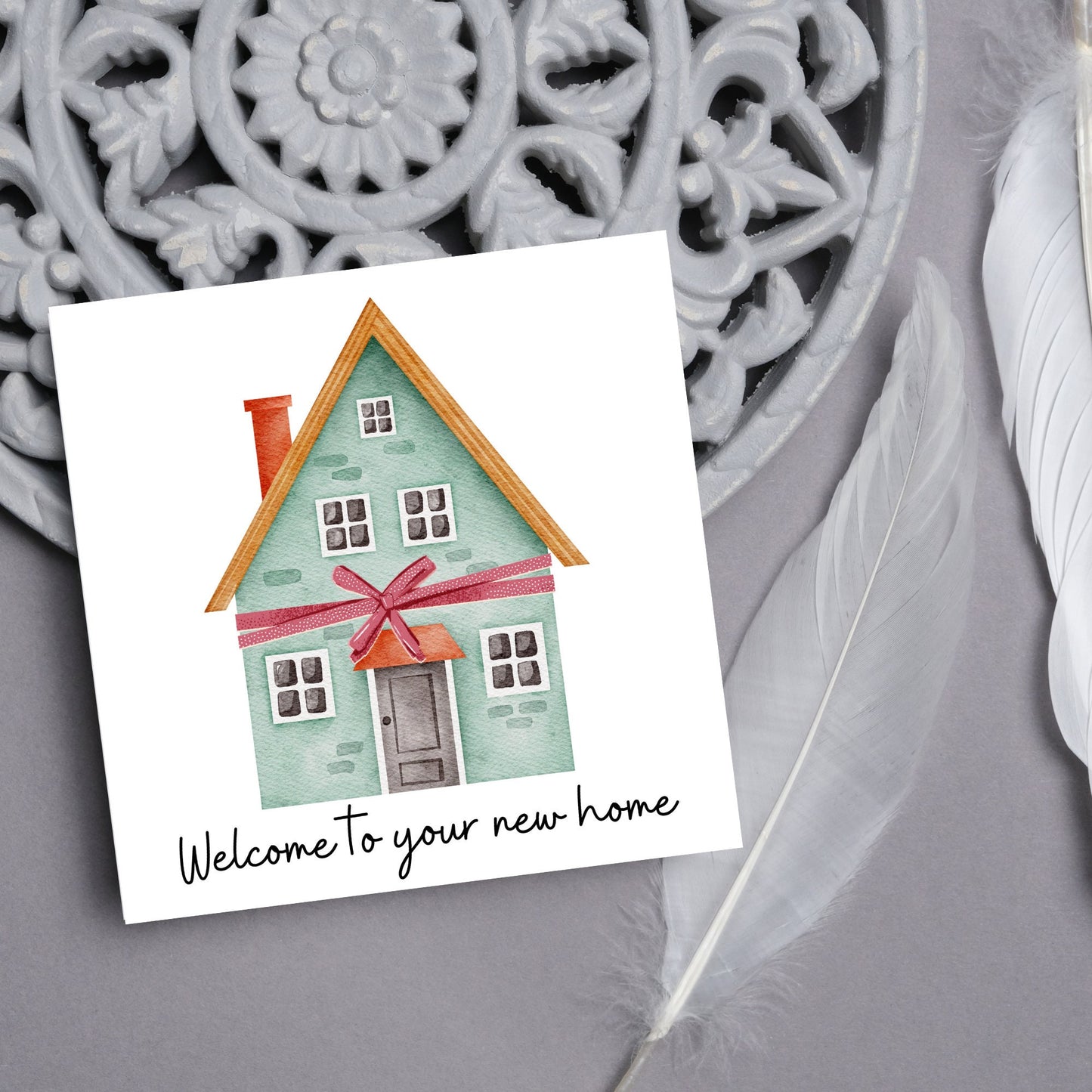Square card with white background and a watercolour style illlustration of a green three storey house with a red bow wrapped around it. Underneath the house ina script font are the words 'welcome to your new home'