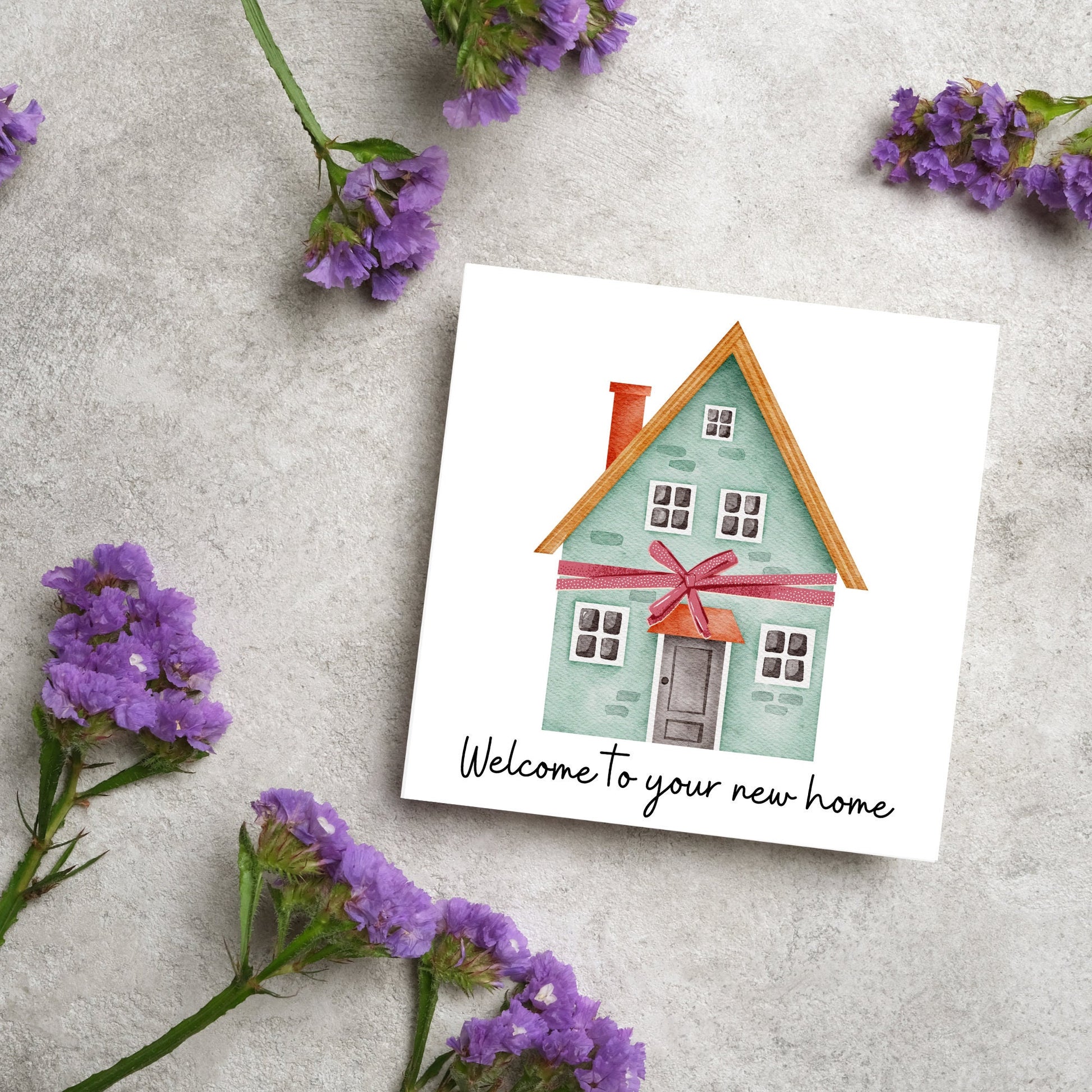 Square card with white background and a watercolour style illlustration of a green three storey house with a red bow wrapped around it. Underneath the house ina script font are the words 'welcome to your new home'