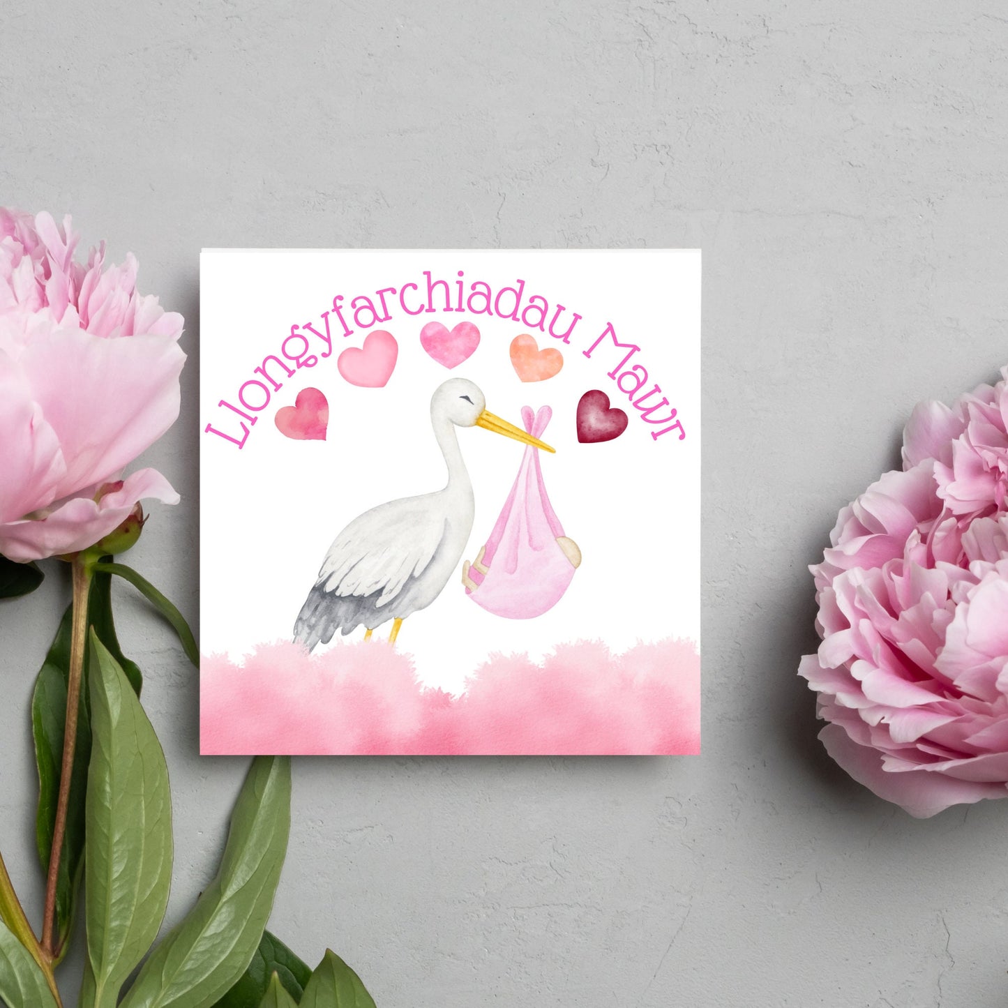 Image of a square greetings card with a stork delivering a baby in a pink blanket. There are 5 pink watercolour hearts above the stork and the at the top is pink text 'llongyfarchiadau Mawr' which translates from welsh as many congratulations
