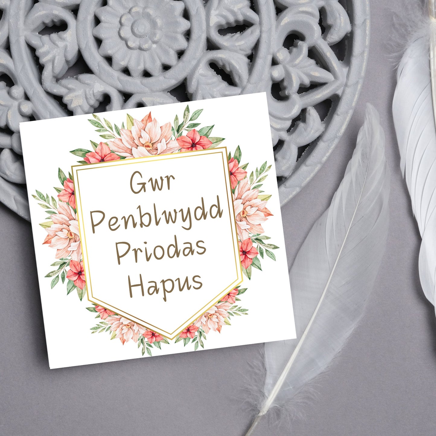 Image of a square wedding anniversary card with 'gwr penblwydd priodas hapus' in a subtle handwritten style font, which translates to 'happy wedding anniversary' in welsh. the text is against a white banner surrounded by pink flowers